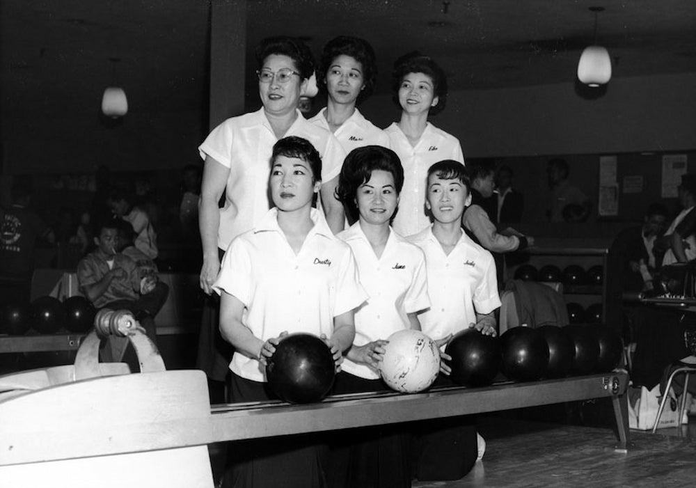Six Japanese American women pose in indoor bowling alley. Three kneel in front and three stand behind, all wearing matching white, collared shirts.
