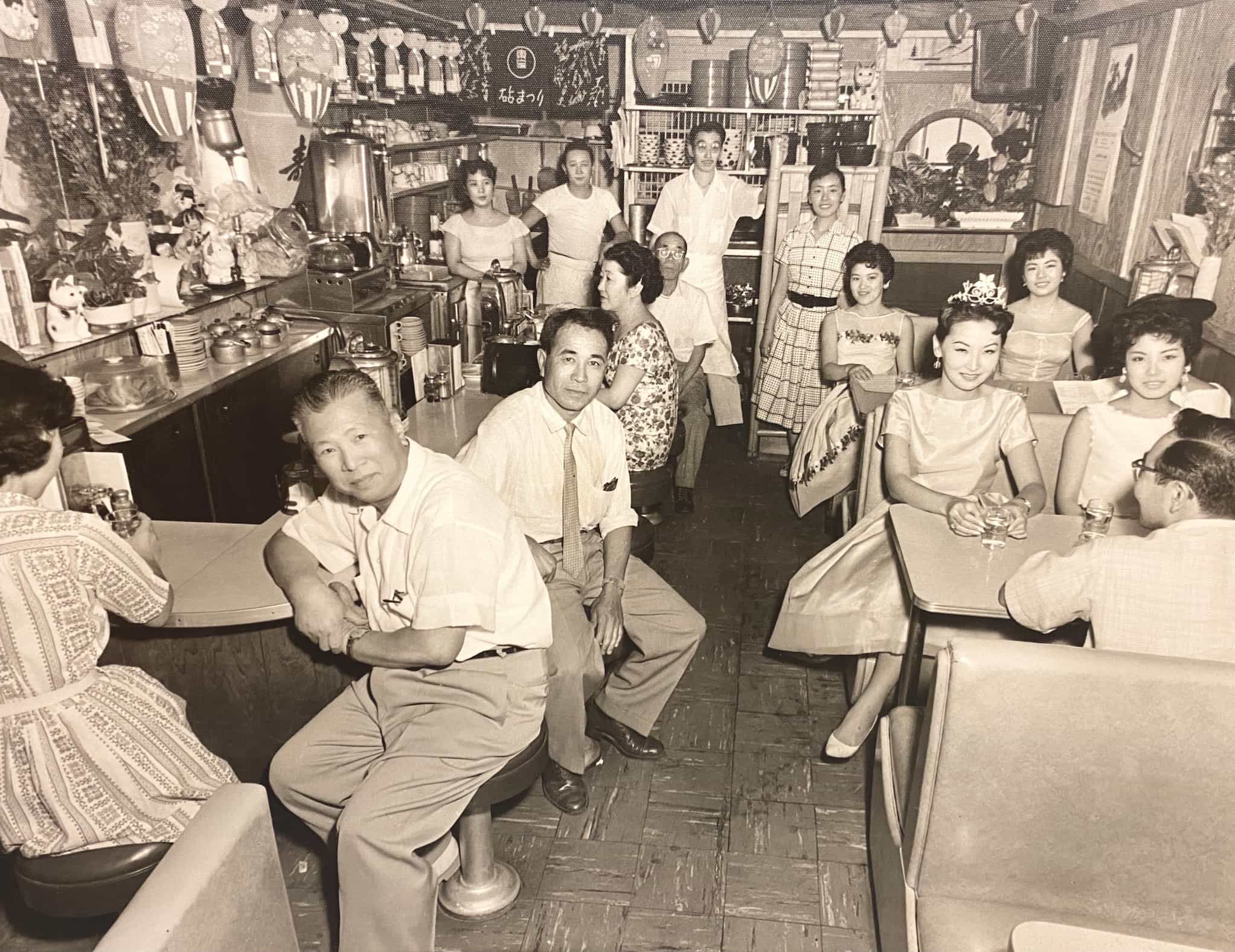 Japanese Americans sit at various tables in restaurant. Some sit at counter top to the left while others sit in booths. Workers stand near the back.