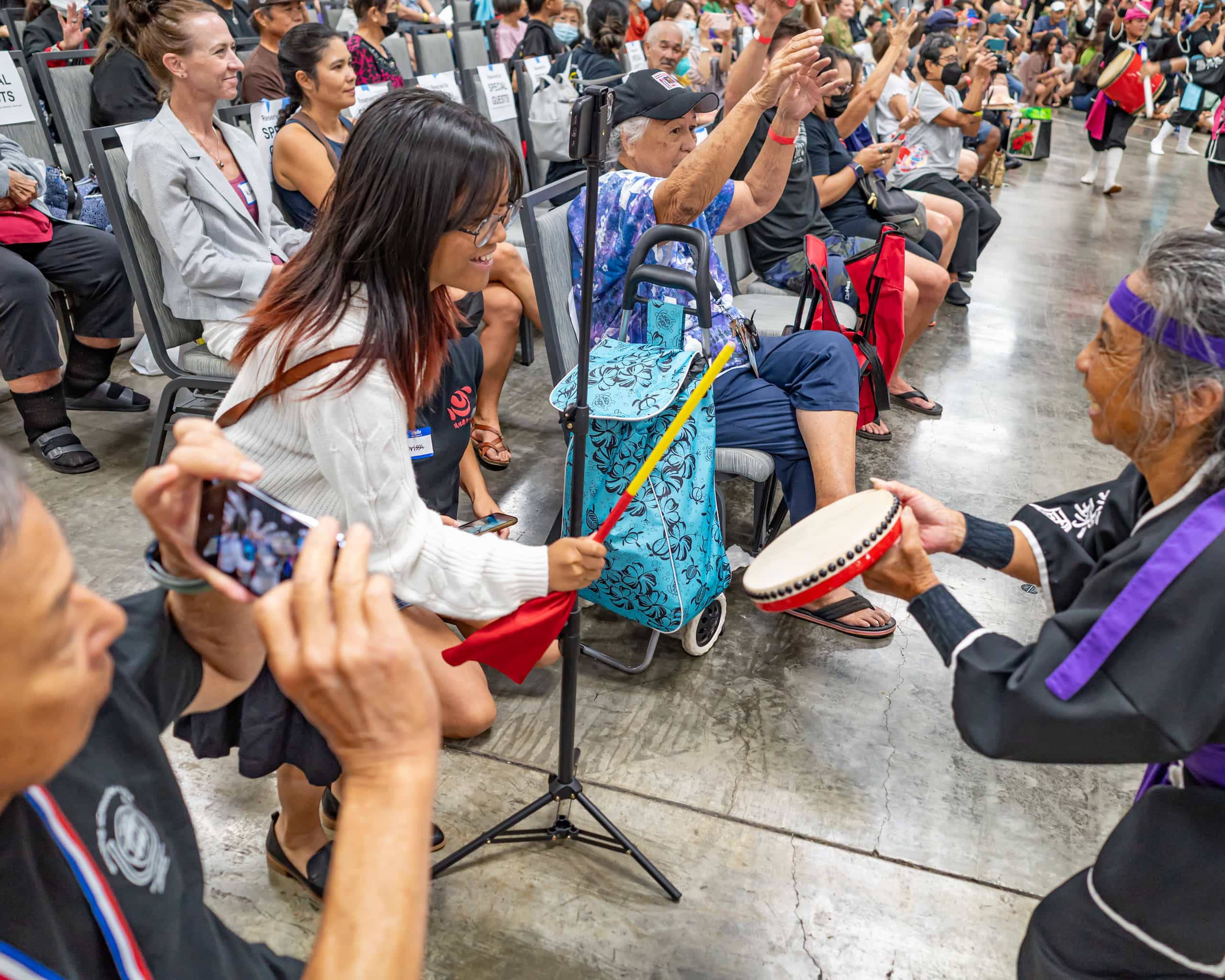 A woman happily strikes a drum held by a elderly person standing before her. A large crowd sits in rows behind her.