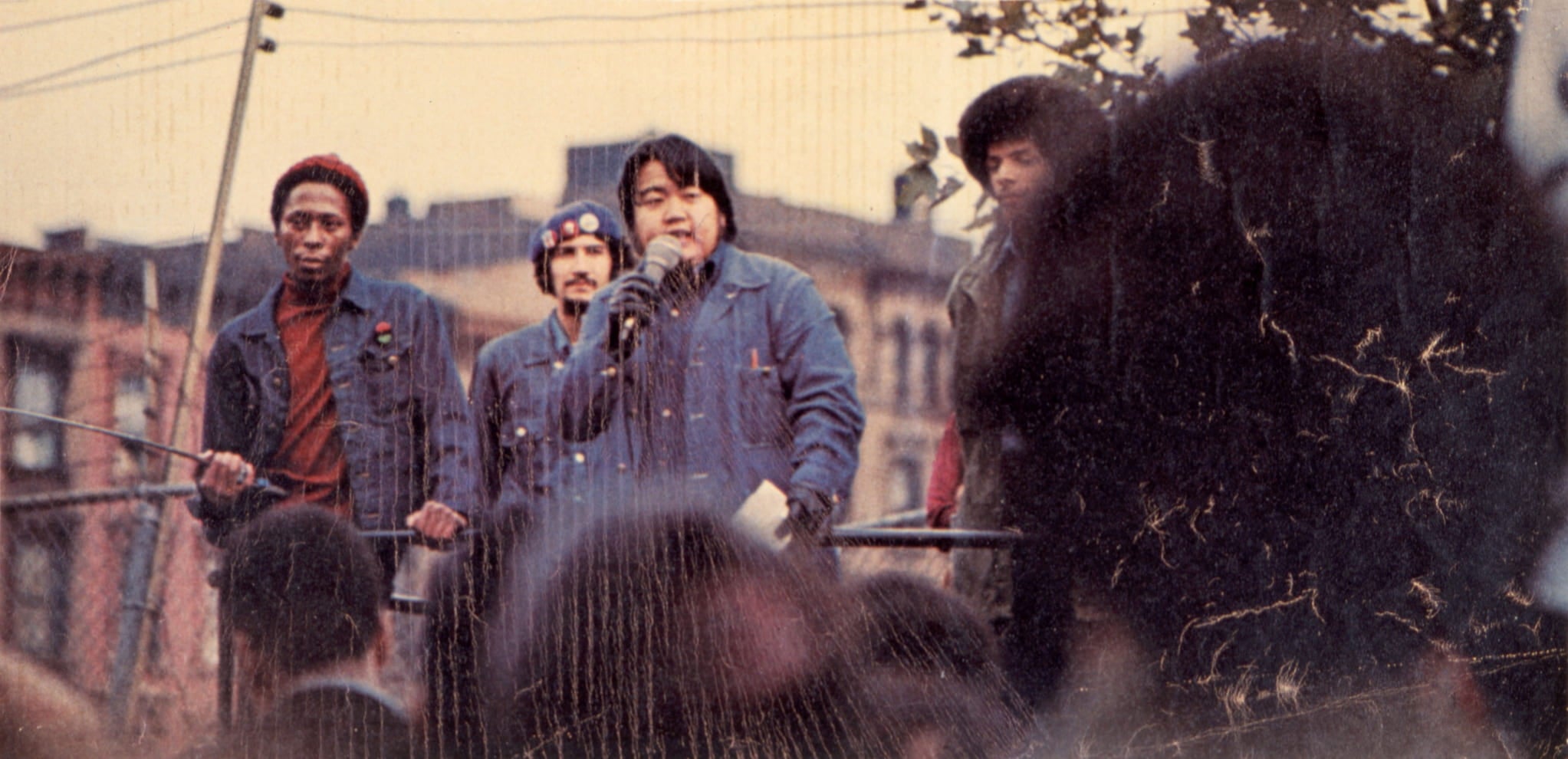 Japanese American activist Warren Furutani holds microphone as he addresses crowd alongside three other men during outdoor rally.