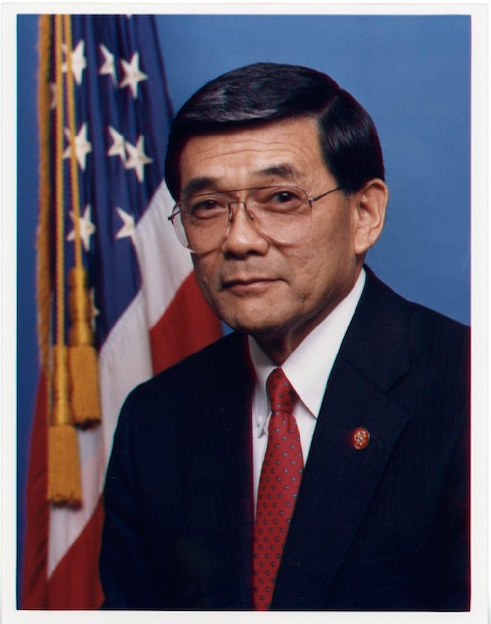 Portrait of Japanese American Norman Mineta. Wearing a black suit and red tie, he sits against blue backdrop with U.S. flag behind him.