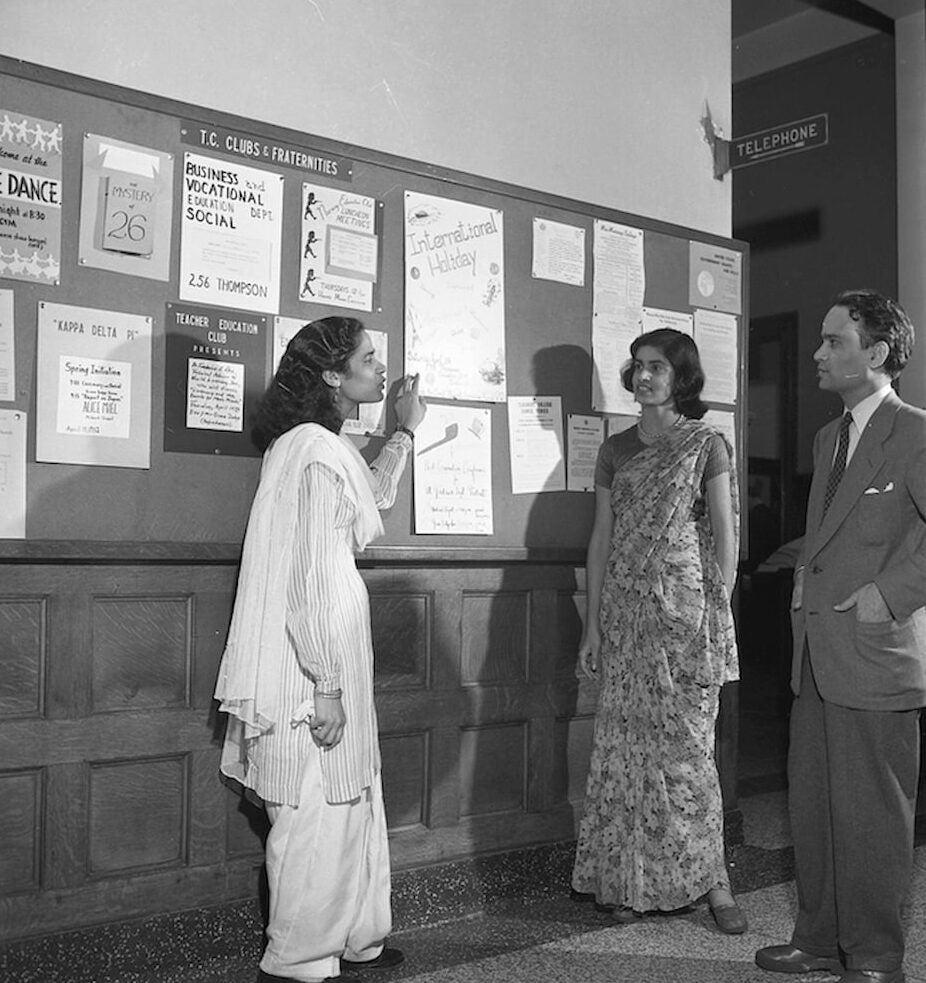 Three Pakistani students talk near bulletin board. A female student, wearing a knee-length tunic and pants, points to pinned flyer on board.