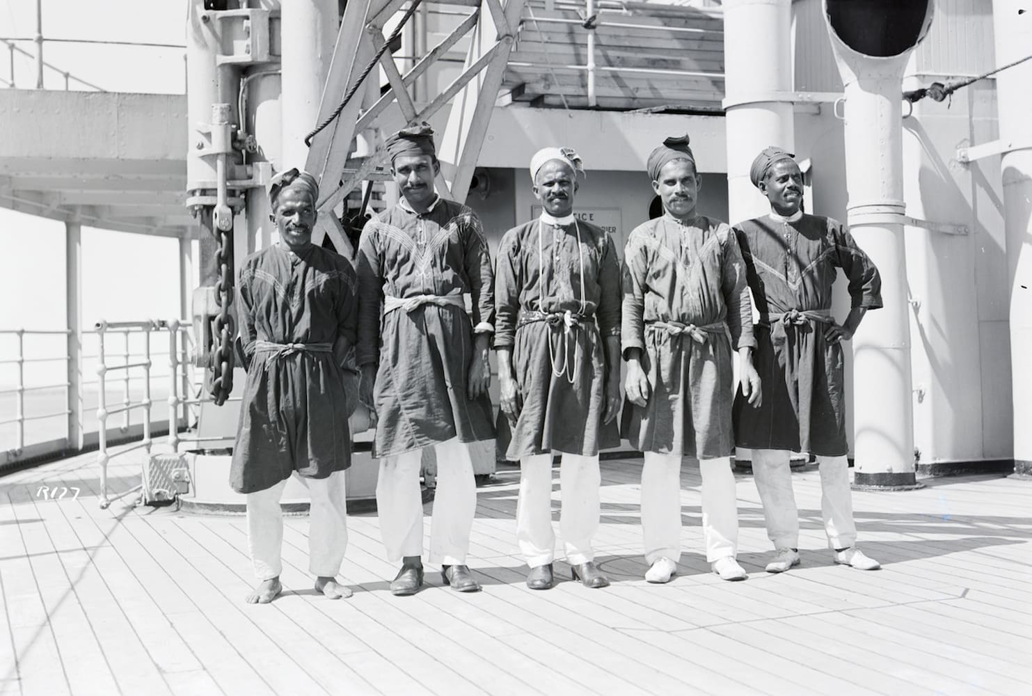 Five South Asian sailors stand in a row on ship's deck. The men wear a uniform of white pants, dark, knee-length tunics, and head coverings.