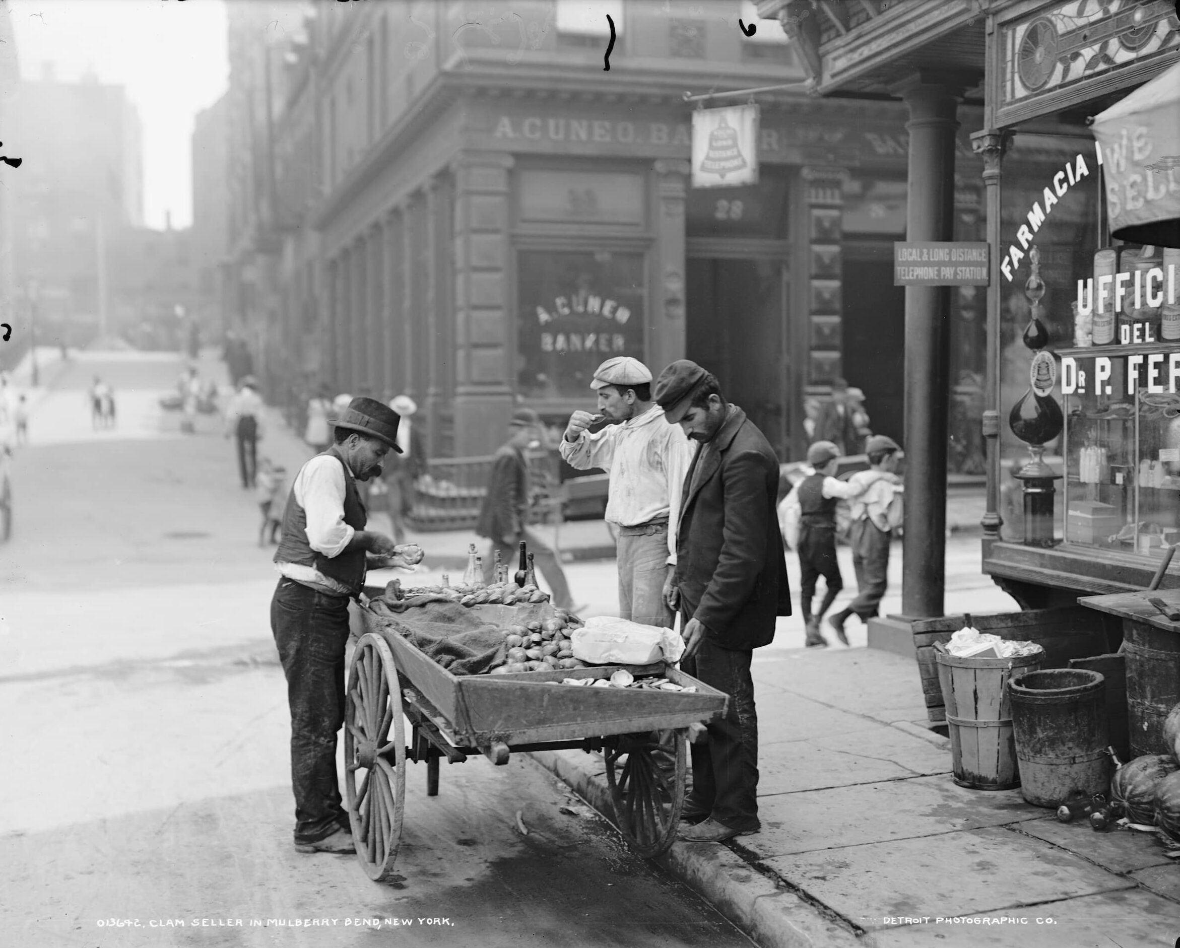 Street vendor in New York shucks and sells clams from a wooden cart. Two men stand opposite, one eating a clam while the other looks down at the cart.