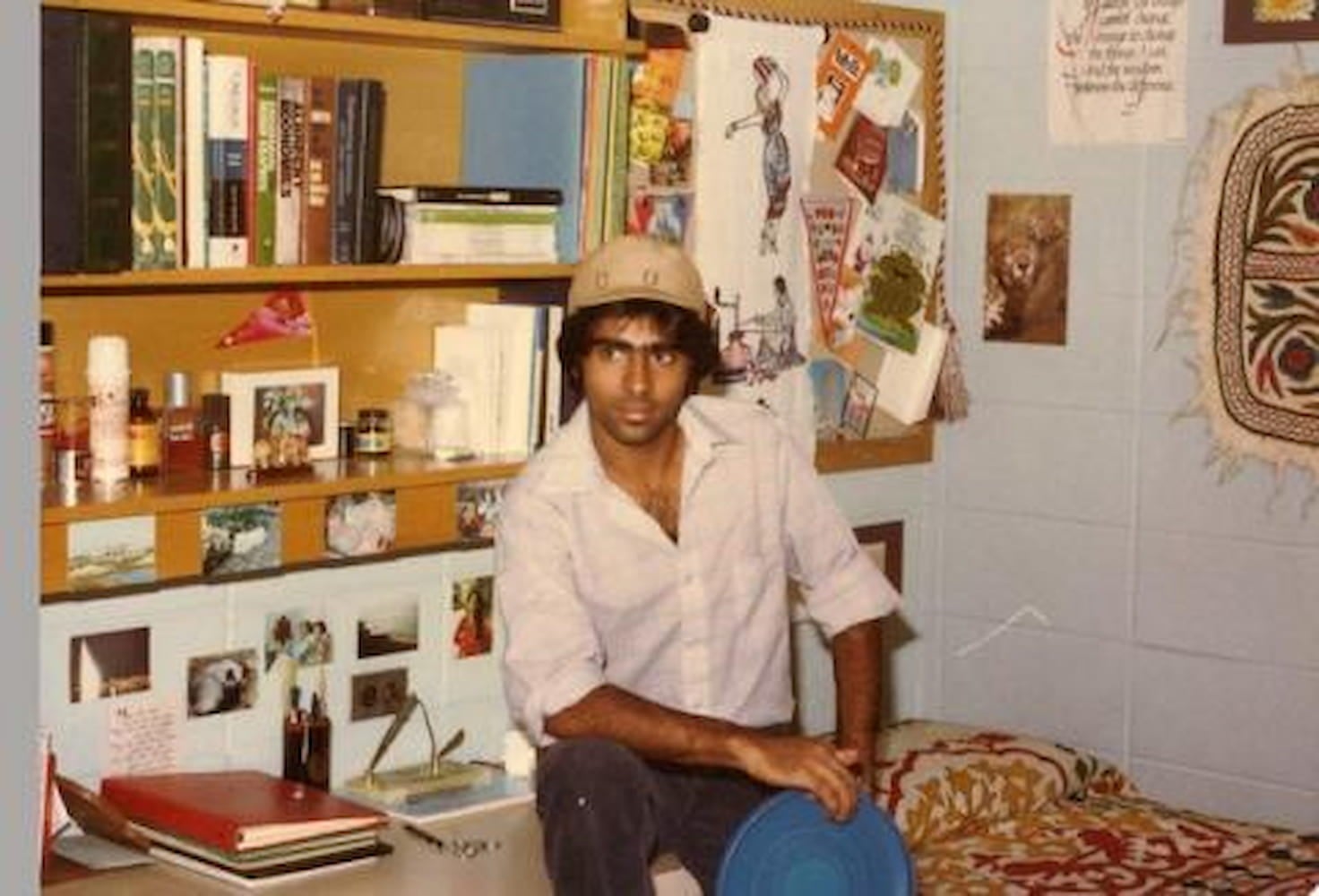 Ali Khataw, wearing a white button down shirt and a tan hat, sits on desk in dorm room. Desk and shelf are stacked with books. Photos cover walls.