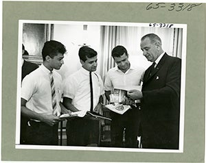President Johnson stands beside three male students from Pakistan and India during White House tour. Each holds booklets as the President flips pages.
