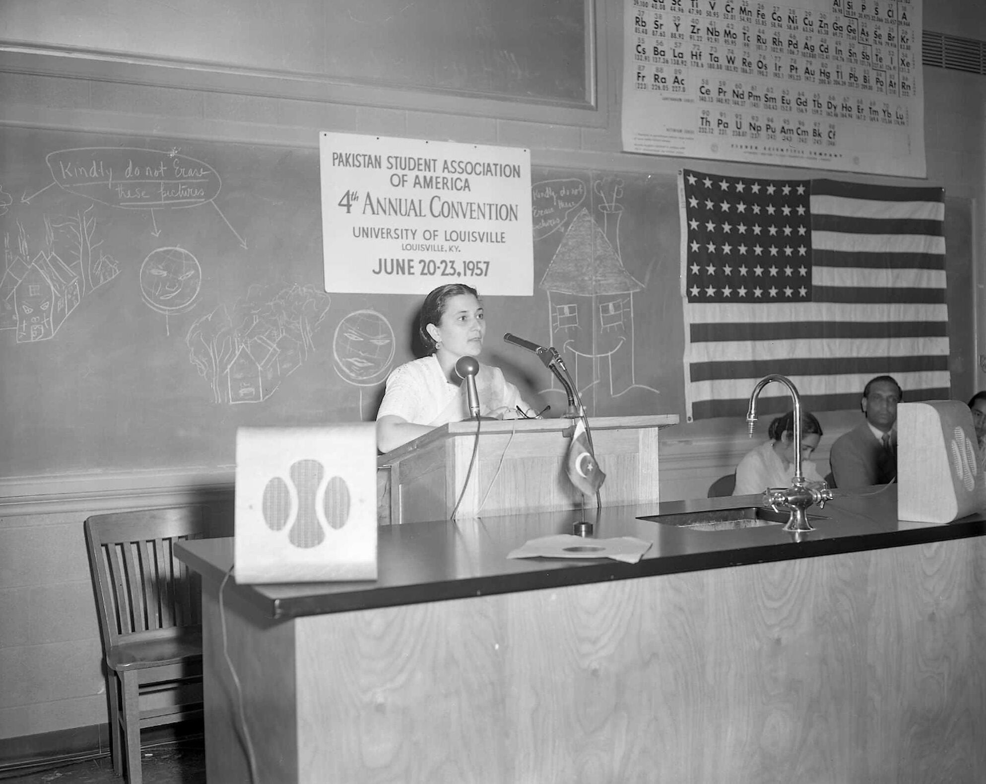 Pakistani woman speaks at classroom podium. Behind her is a sign that reads, "Pakistan Student Association of America" and an American flag.
