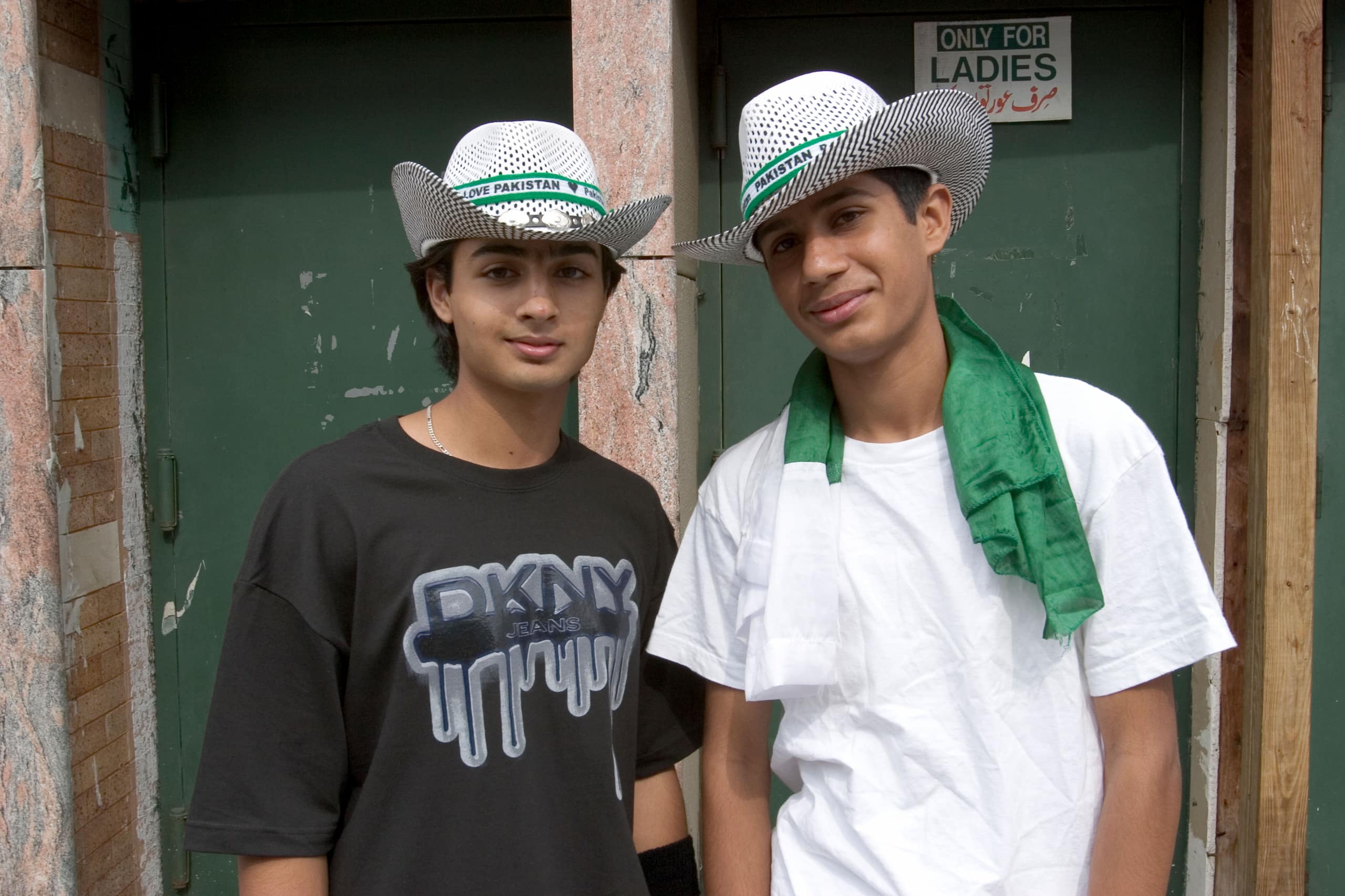Two young men stand next to each other in front of green doors. Both wear white western style hats with ribbons that reads Pakistan around the base.
