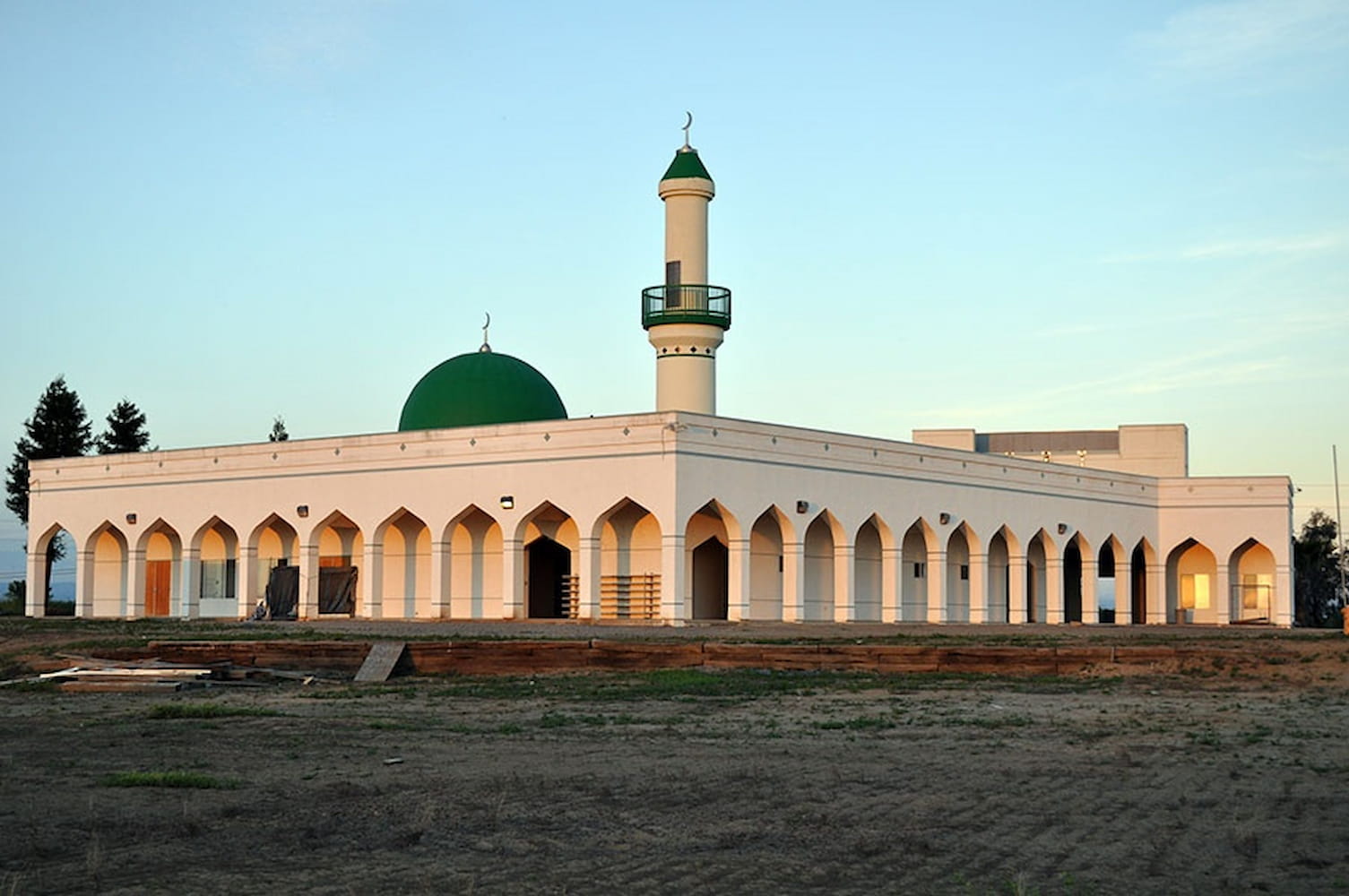 Corner view of mosque on large grassy lot. Above the mosque's columns and arches is a green dome and minaret.