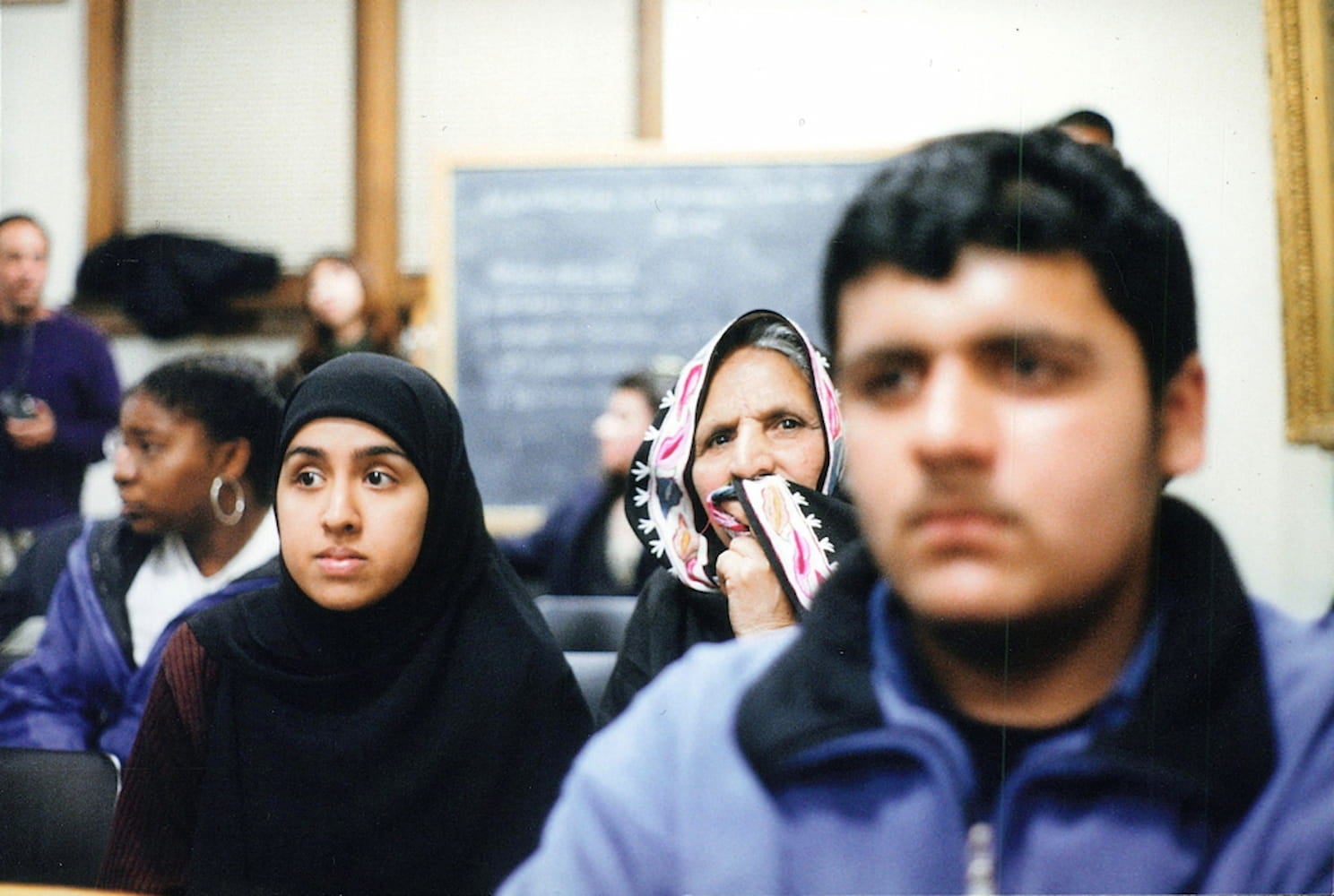 Two women in headscarves sit in second row during community forum. Unfocused in the foreground, a man in blue jacket sits in front of them.