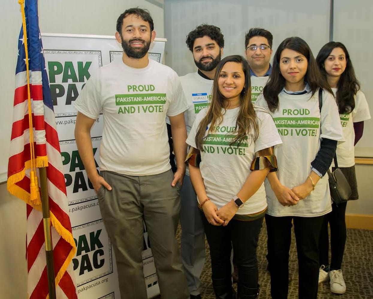 Young volunteers, all wearing "Proud Pakistani-American And I Vote" shirts, stand next to an American flag inside an office and smile for photo.