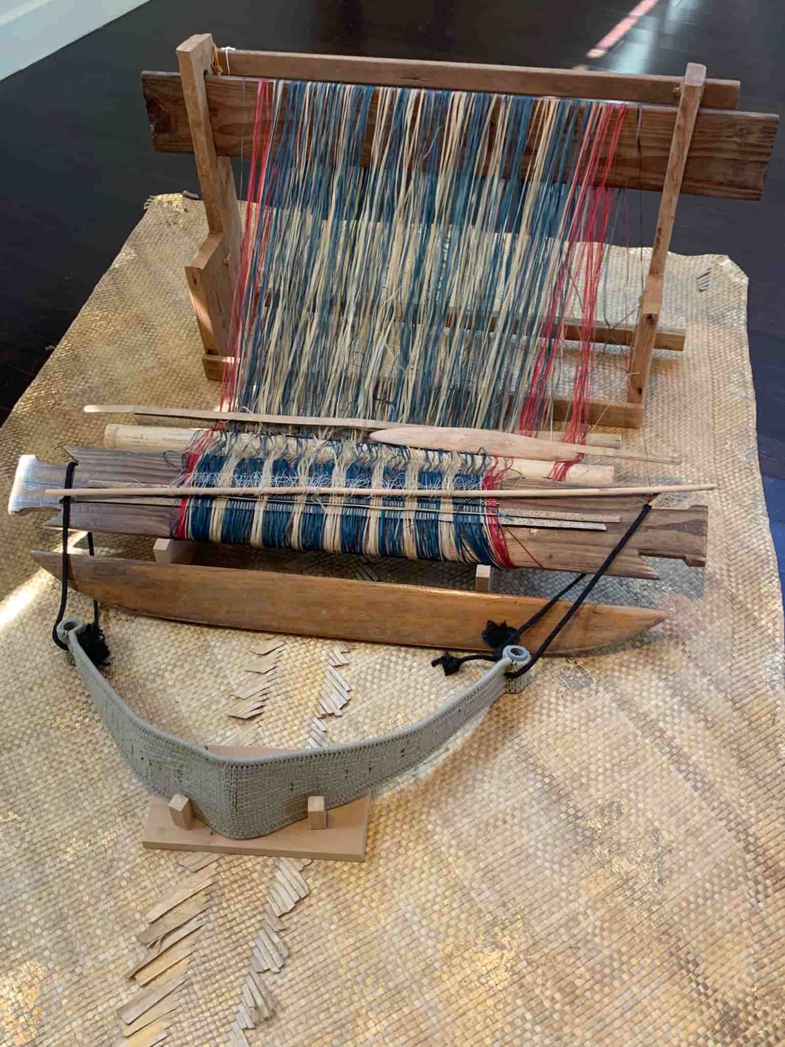 A wooden loom, with a gray strap attached for weavers to wrap around their backs, is on woven mat. Red, blue, and natural fibers are being woven.