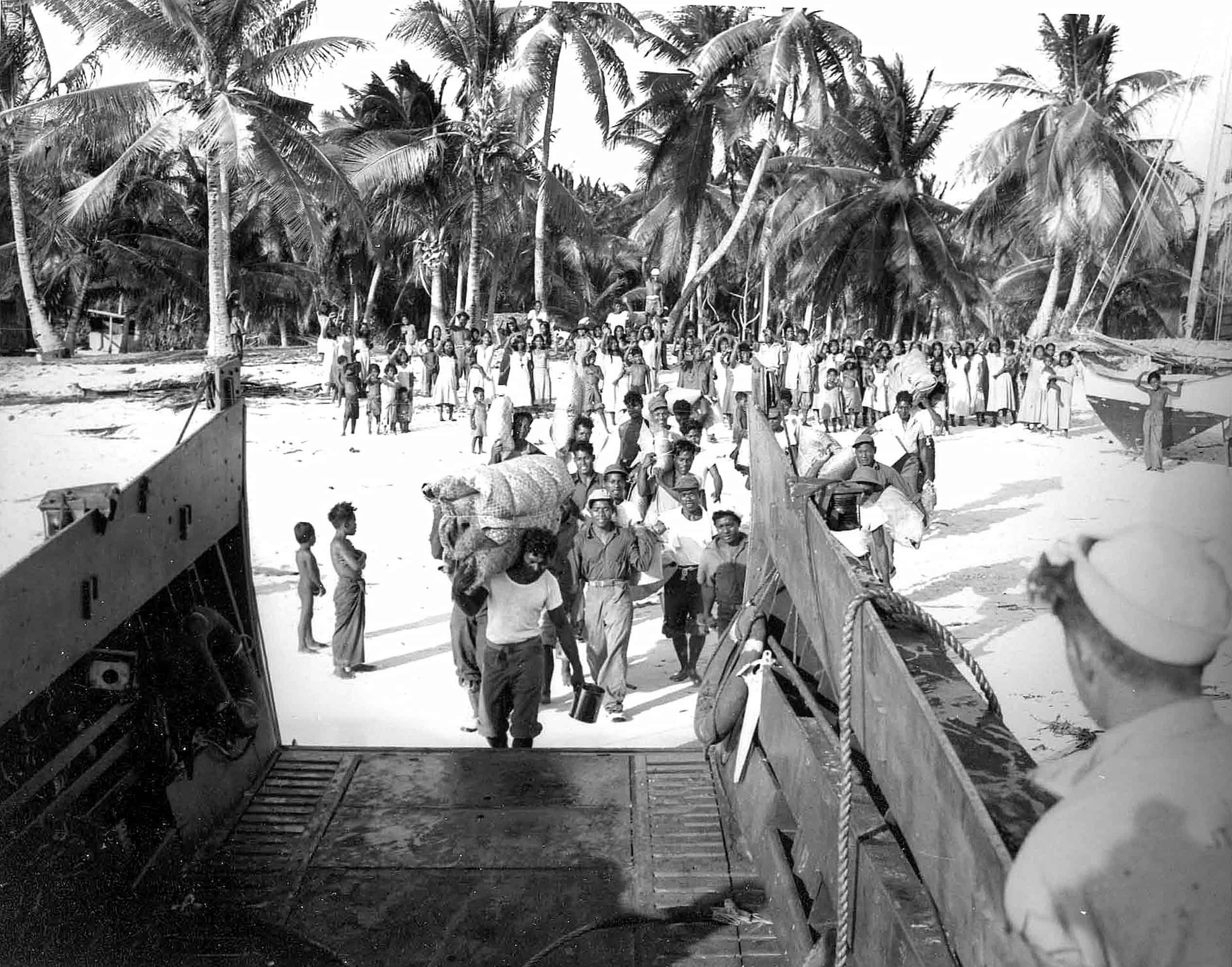 From the point of view of a naval landing craft, a group walks across the beach with their belongings. Behind them, a crowd stands on sand and waves.