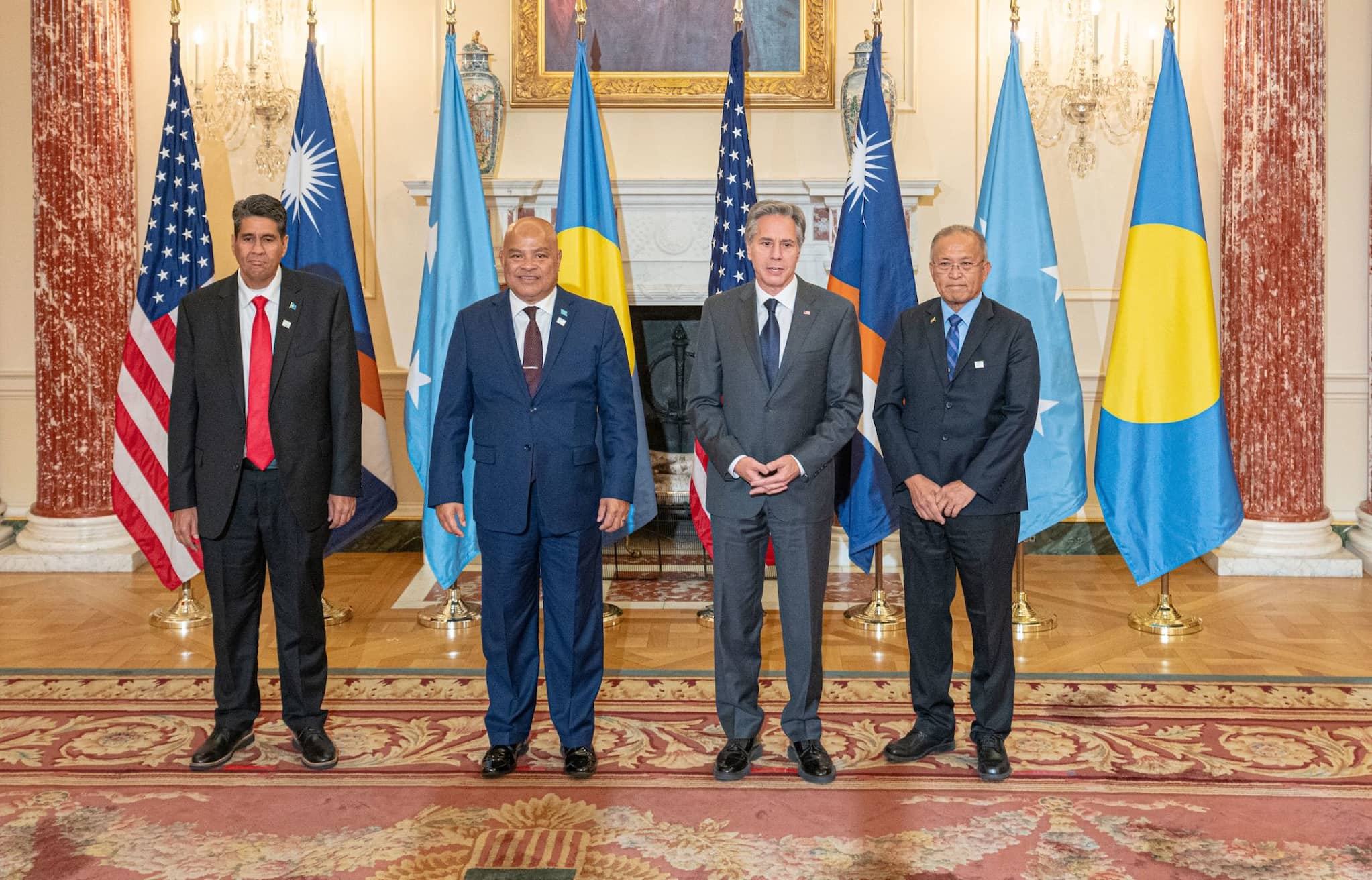 Four men in suits stand in front of flags from the U.S., Republic of the Marshall Islands, Federated States of Micronesia, and Republic of Palau.