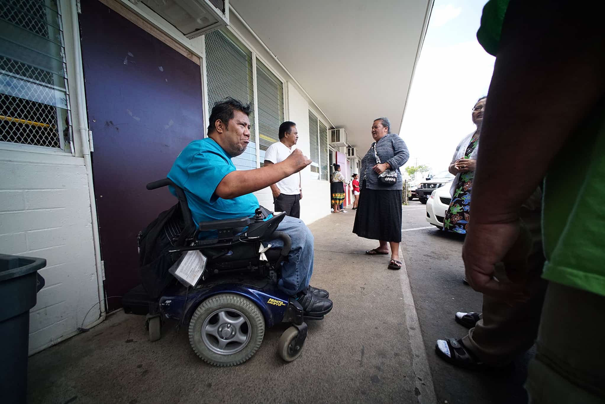 Dr. Joakim “Jojo” Peter, a Micronesian man dressed in bright blue who uses a motorized wheelchair, addresses people standing around him on outdoor walkway.