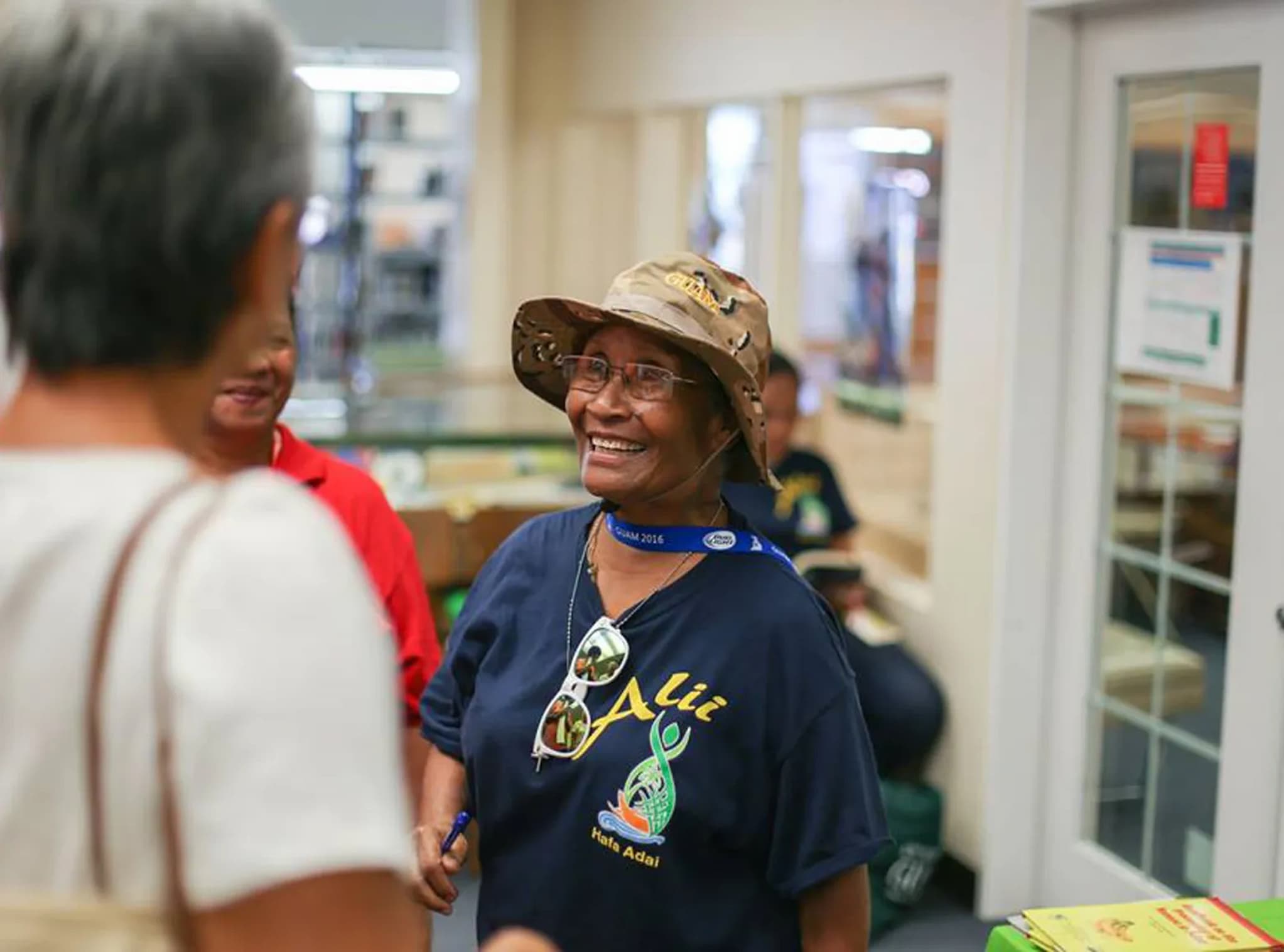 Hermana Remarui, a Palauan woman wearing a boonie hat and dark blue t-shirt, smiles and engages with a small group of people at a library.