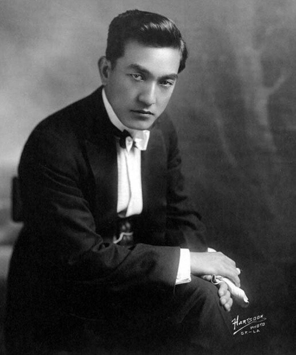 Portrait of Japanese born actor Sessue Hayakawa. Hayakawa, wearing a suit and bowtie, sits before studio backdrop with his hands resting on his knee.