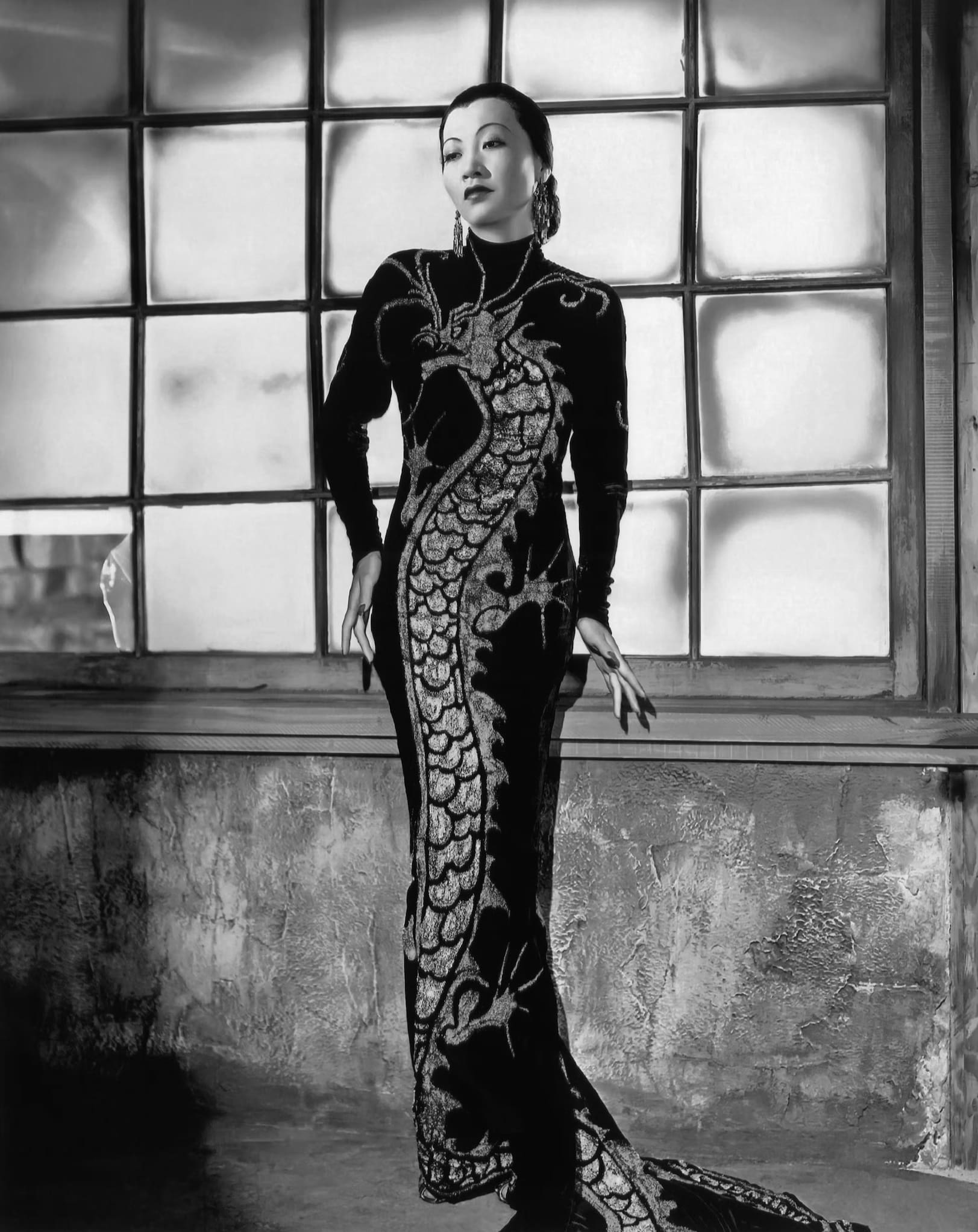 Chinese American actress Anna May Wong, in a long-sleeved dress with an embroidered dragon covering the front, poses with a large window behind her.