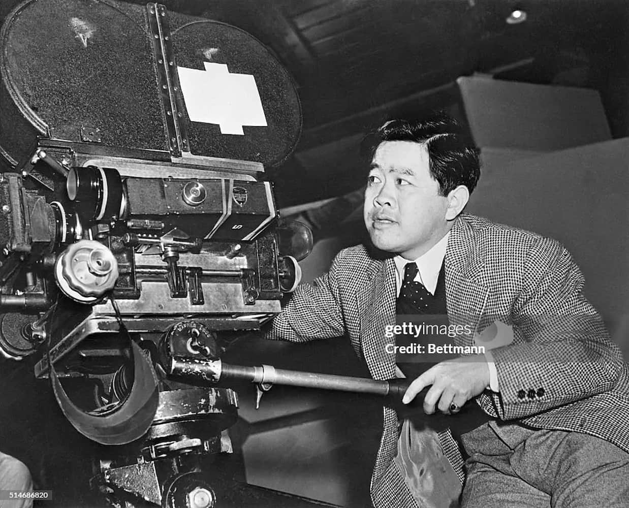 James Wong Howe, a Chinese American cinematographer dressed in a tweed jacket, vest, pants, shirt and tie, sits behind a movie camera.