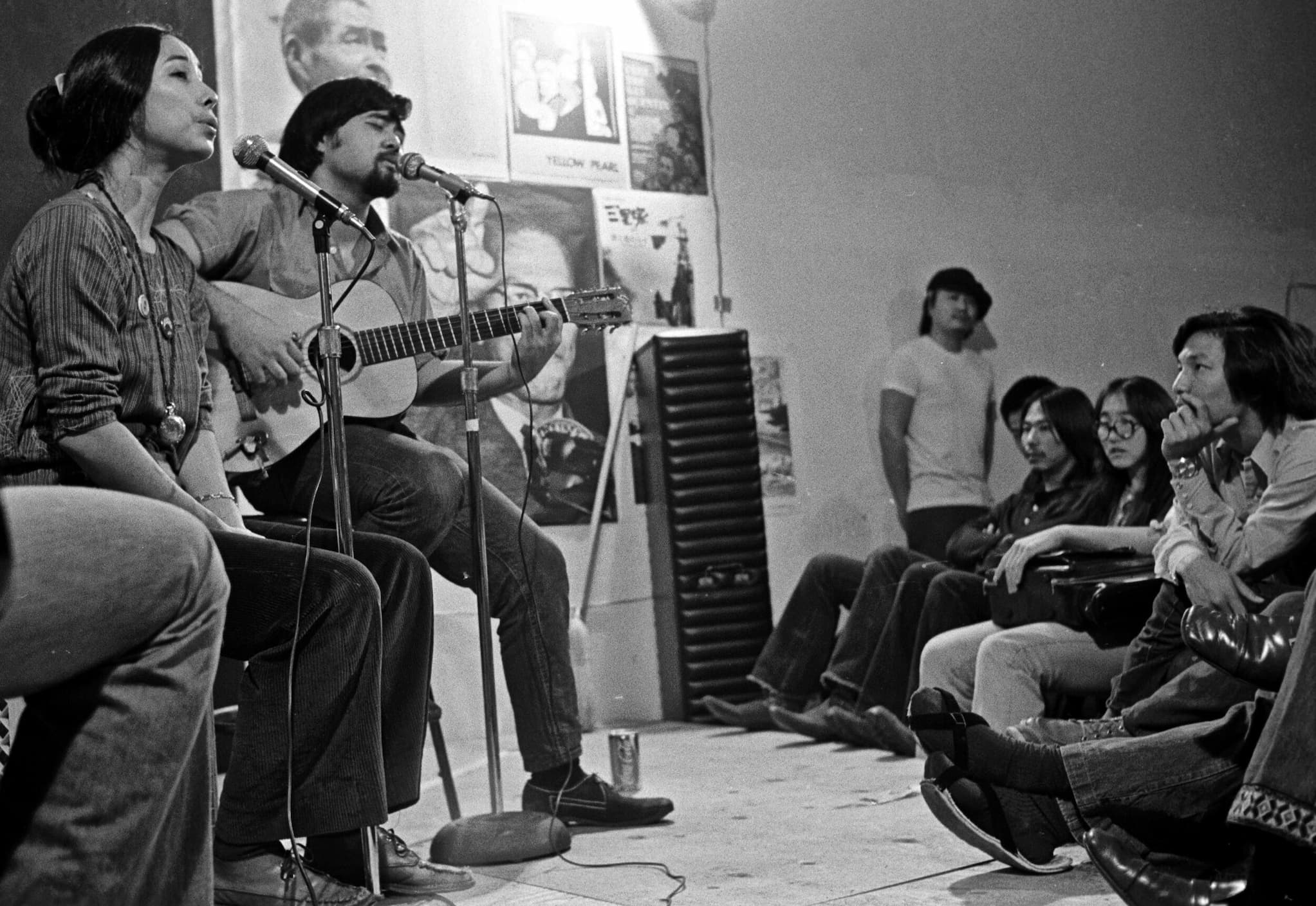 Nobuko Miyamoto, seated before small crowd, sings into microphone while Chris Iijima, seated beside her, sings and plays guitar during a performance.
