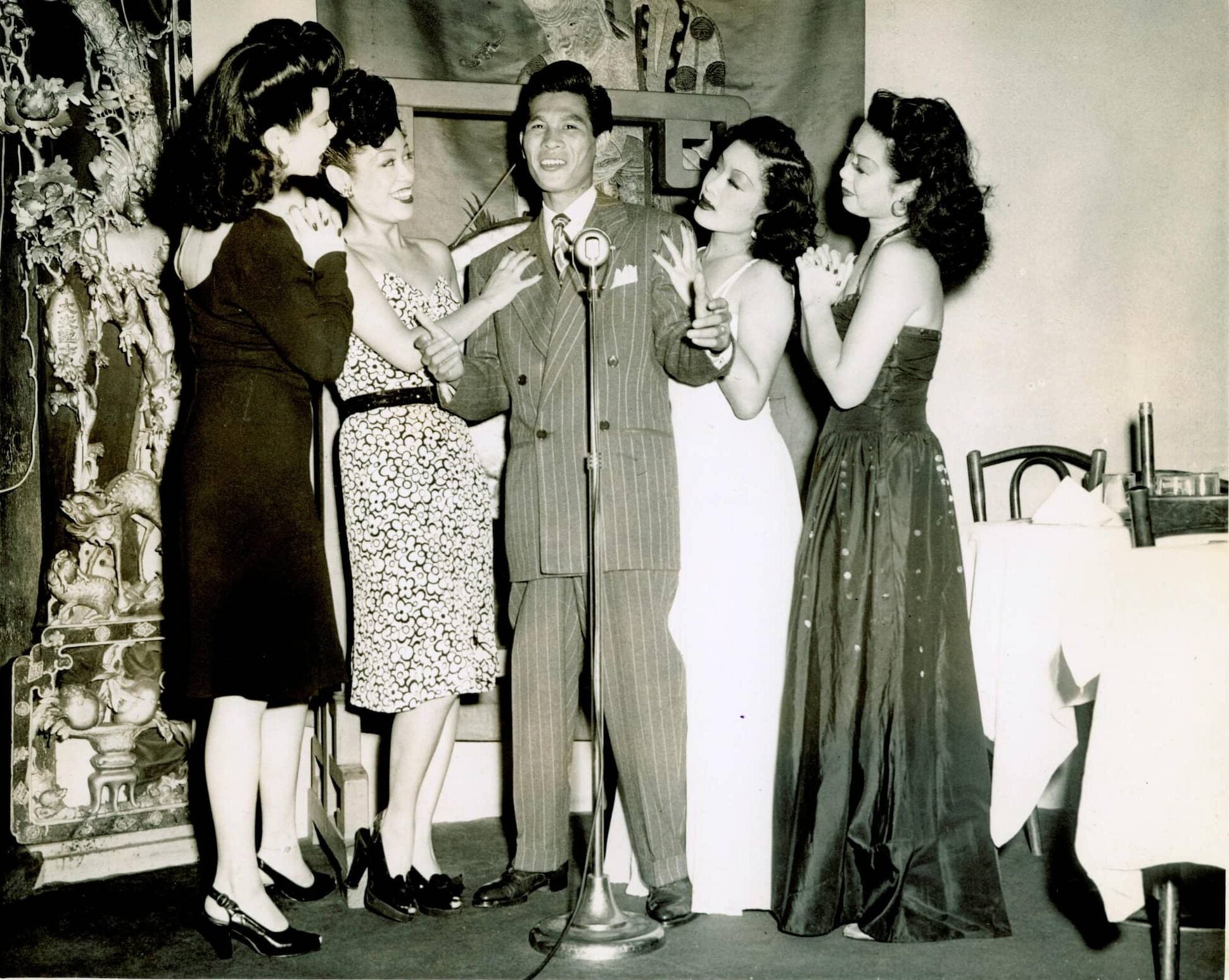 In a nightclub, Larry Ching, dressed in a suit, stands before a microphone. Around him a group of women wearing dresses smile and look towards him.