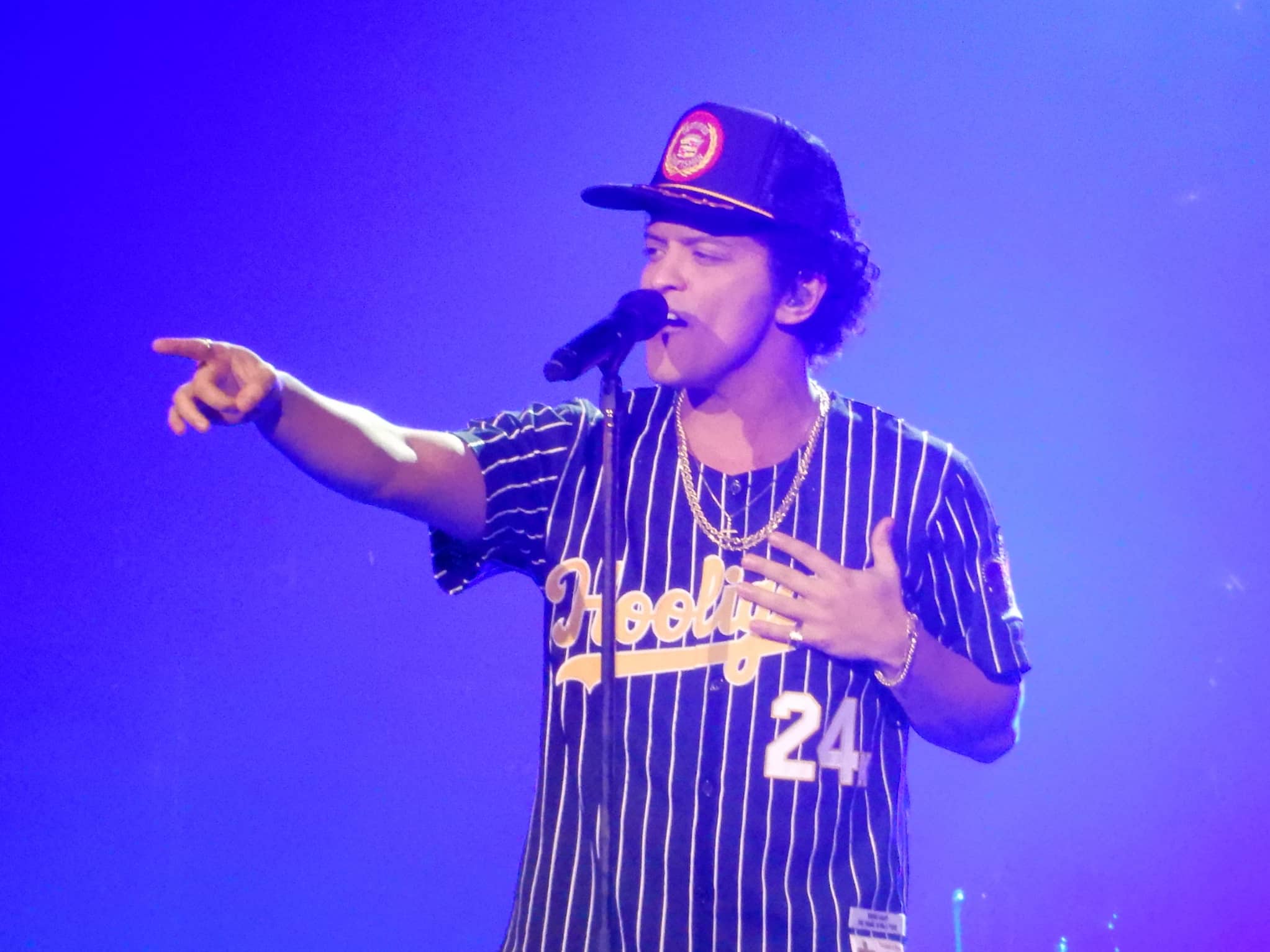 Bruno Mars, wearing a baseball cap and striped jersey, sings on stage. He points to the audience and holds his left hand up to his heart.