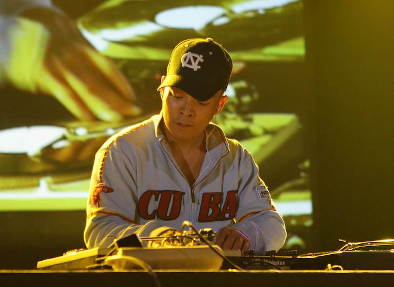 Filipino American DJ Q-Bert, wearing a zip-up hoodie and baseball cap, performs on stage at a festival. Footage of his performance is on display.