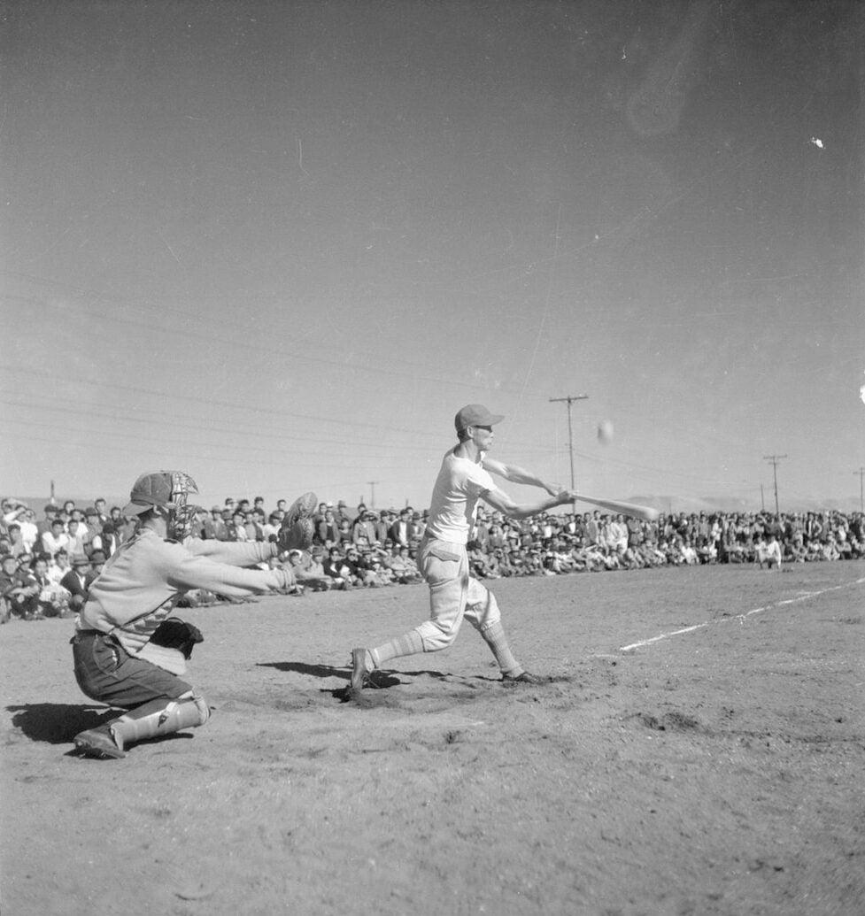 Japanese American man swings bat towards baseball. Pitcher crouches behind him. A crowd lines perimeter of baseball field.