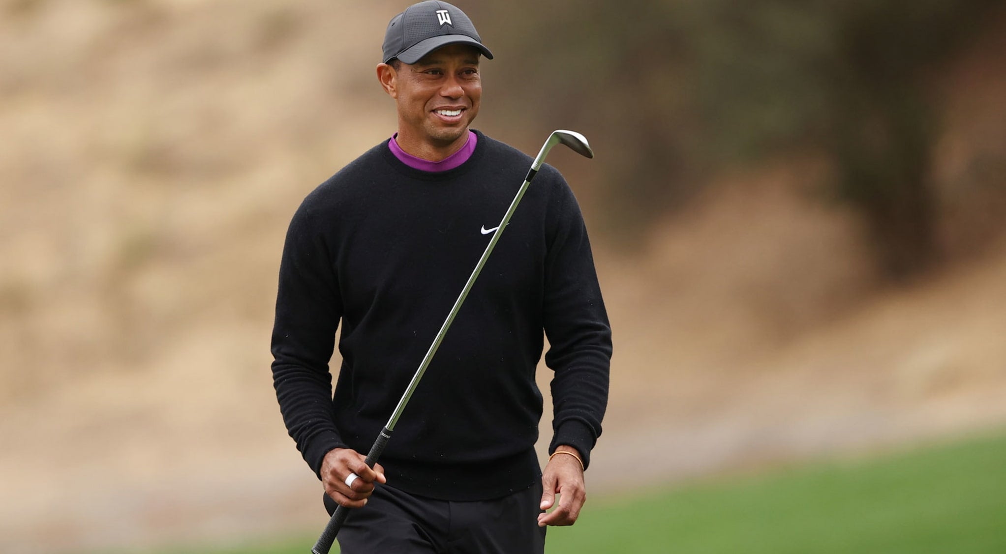 Tiger Woods, wearing a sweatshirt and golf cap, carries a golf club in one hand and smiles as he walks on the green.