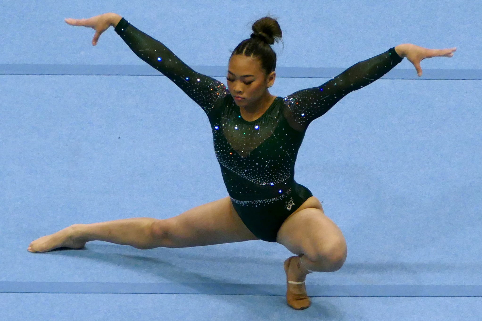 Gymnast Sunisa Lee, dressed in dark, long-sleeved leotard, performs floor routine. Lee squats with one leg extended straight and arms raised.