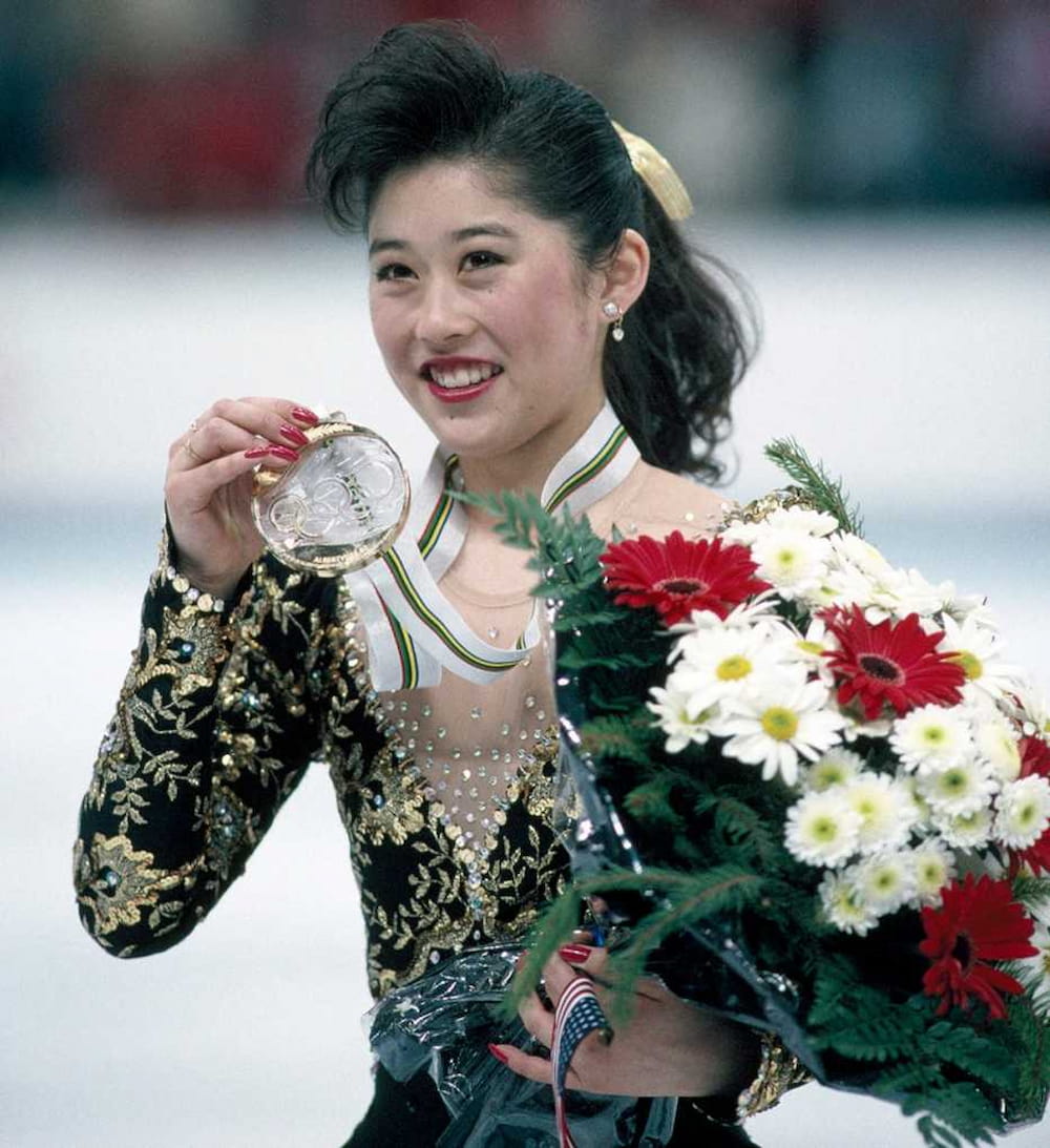 Kristi Yamaguchi, dressed in an embellished figure skating dress, holds a large bouquet in one hand and her Olympic gold medal in the other.