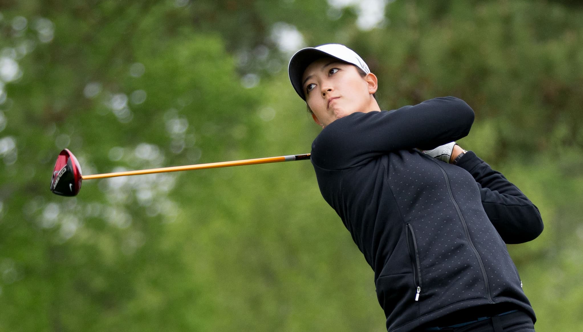 Golfer Michelle Wie, a Korean American woman dressed in a jacket and golf cap, swings a golf club with foliage behind her.