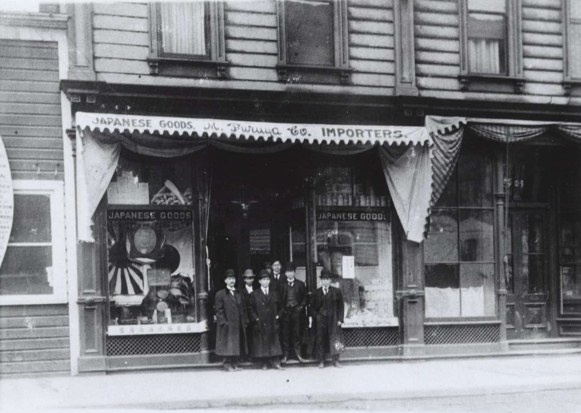 Seven Japanese American men in three piece suits, trench coats, and hats, stand in front of M. Furuya Company. Japanese goods are in window displays.