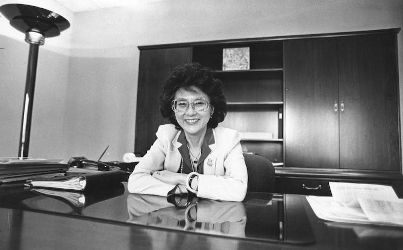 Betty Tom Chu, wearing a suit and glasses, smiles at camera as she sits with her arms folded on desk in front of her. A large credenza is behind her.