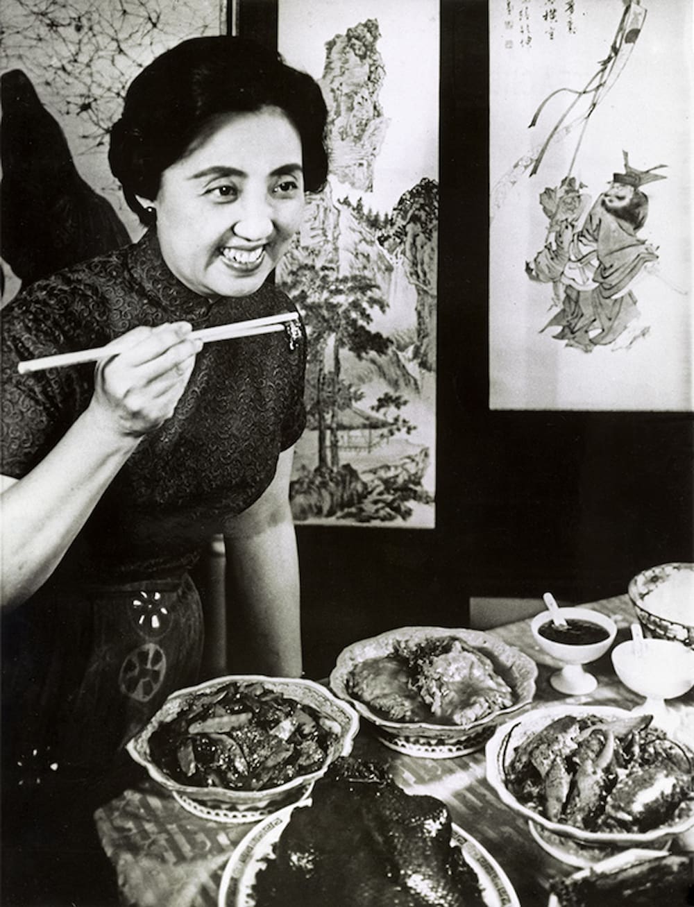 Chinese American chef Joyce Chen holds chopsticks and samples her food while smiling. Multiple dishes of food are displayed on the table before her.