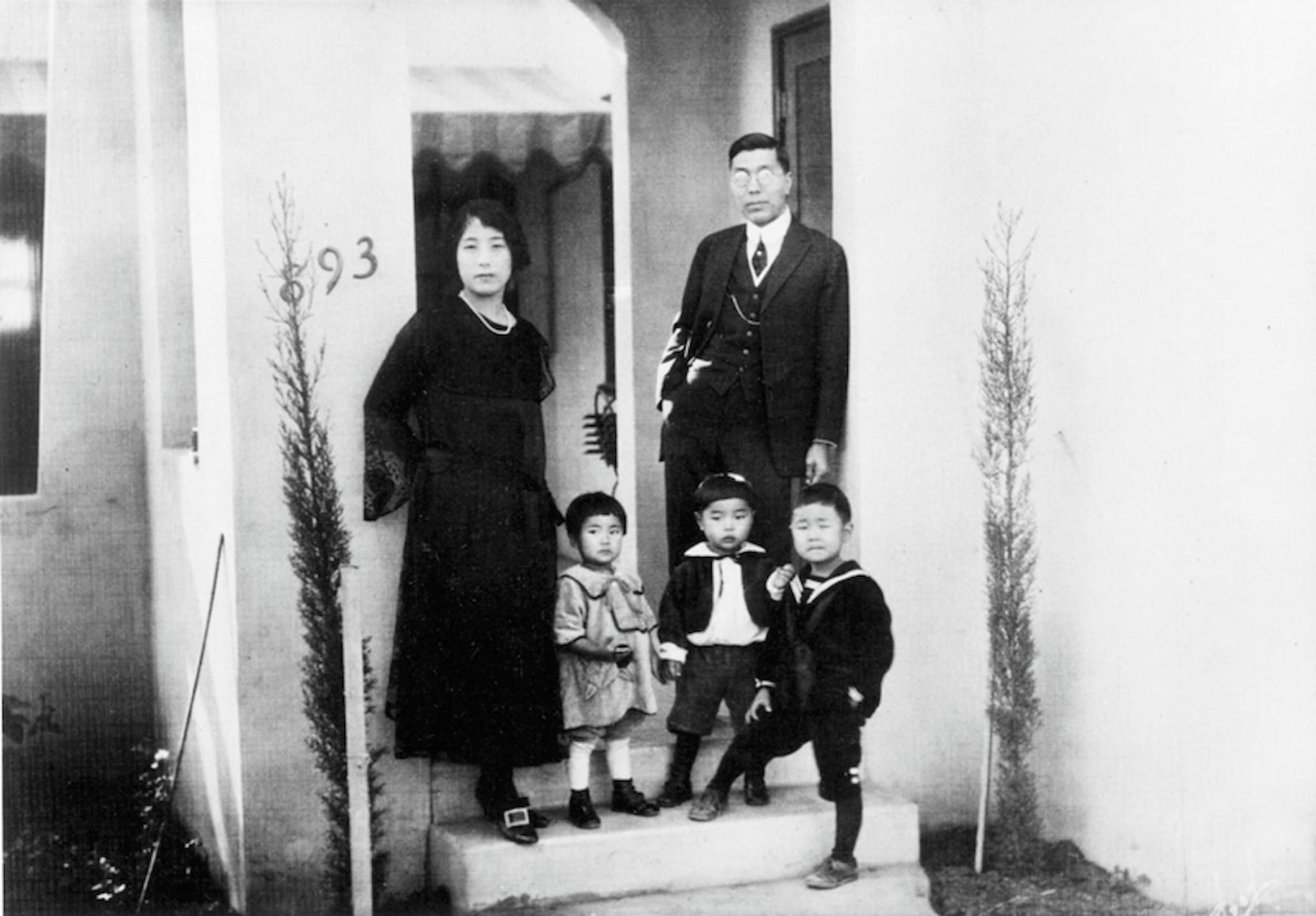 A Japanese husband and wife stand at the doorstep of their home. Their young children, a daughter and two sons, stand beside them on front step.