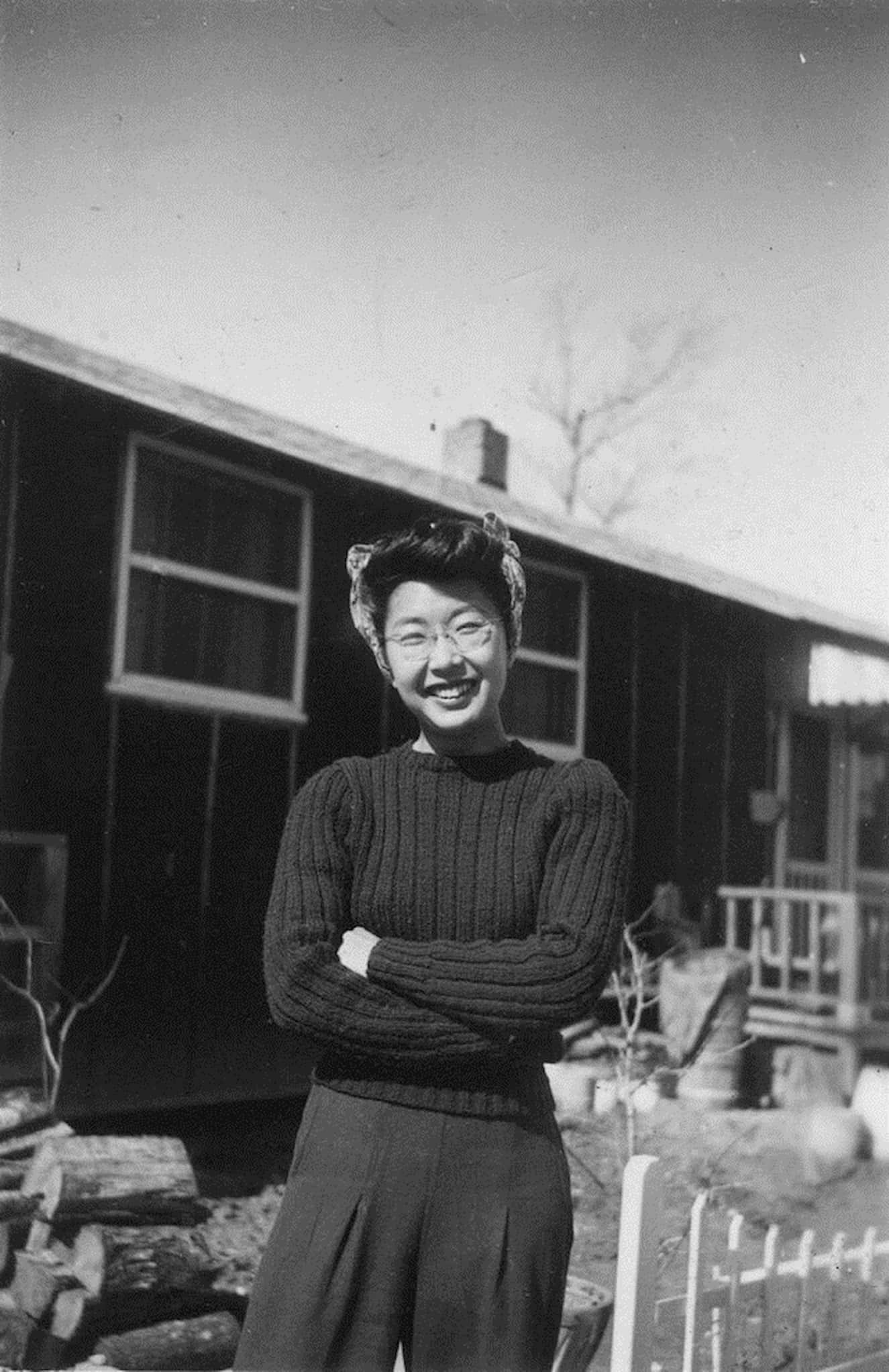 Japanese American Yuri Kochiyama dressed in a wool sweater, slacks and head scarf, smiles with arms crossed in front of the barracks in Santa Anita.