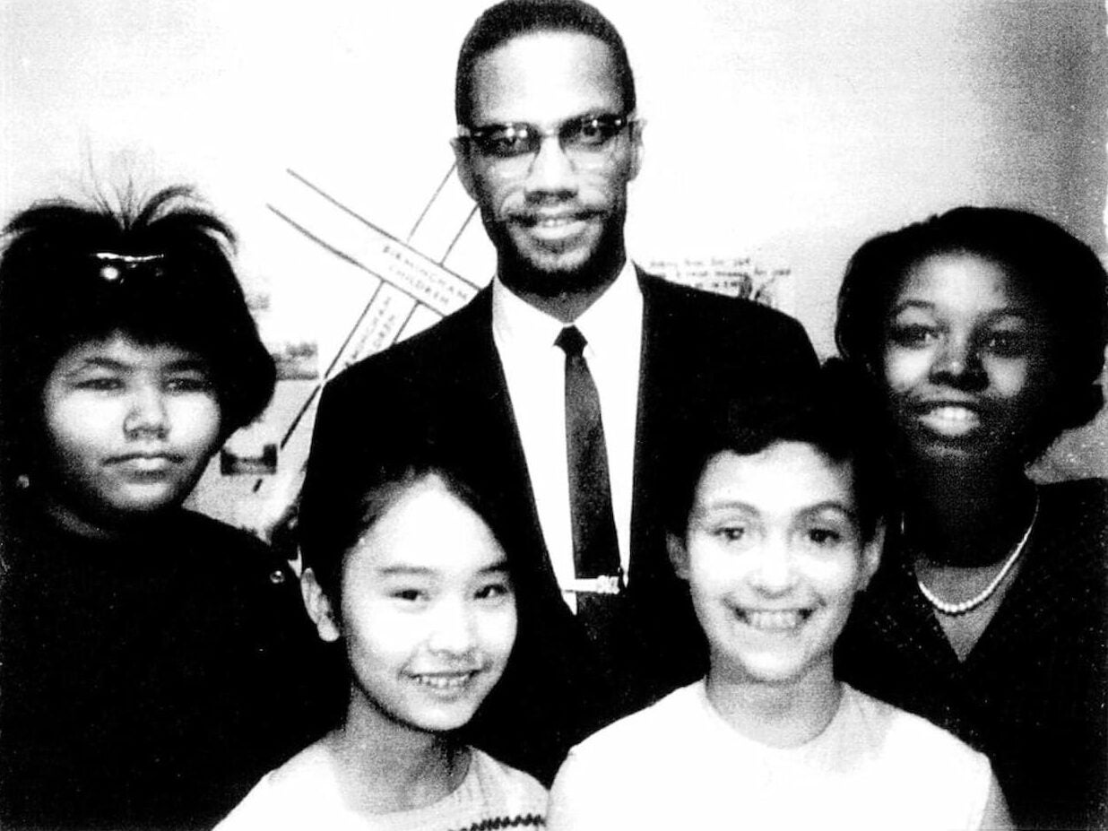 African American activist Malcolm X, in a dark suit and tie, stands at center with four young women of color. Audee Kochiyama stands in front of him.