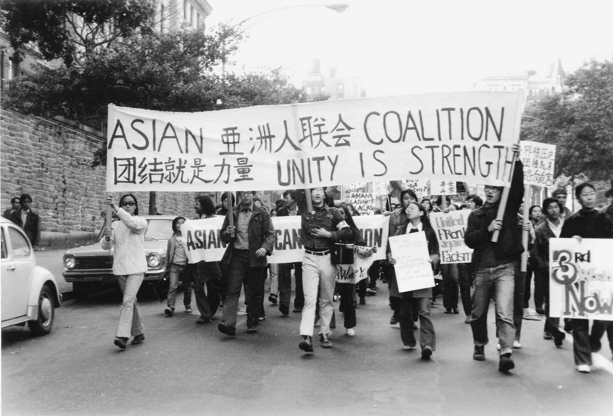 Asian American protestors walk through street holding banners and signs. Banner reads "Asian Coalition Unity is Strength" in English and Mandarin.