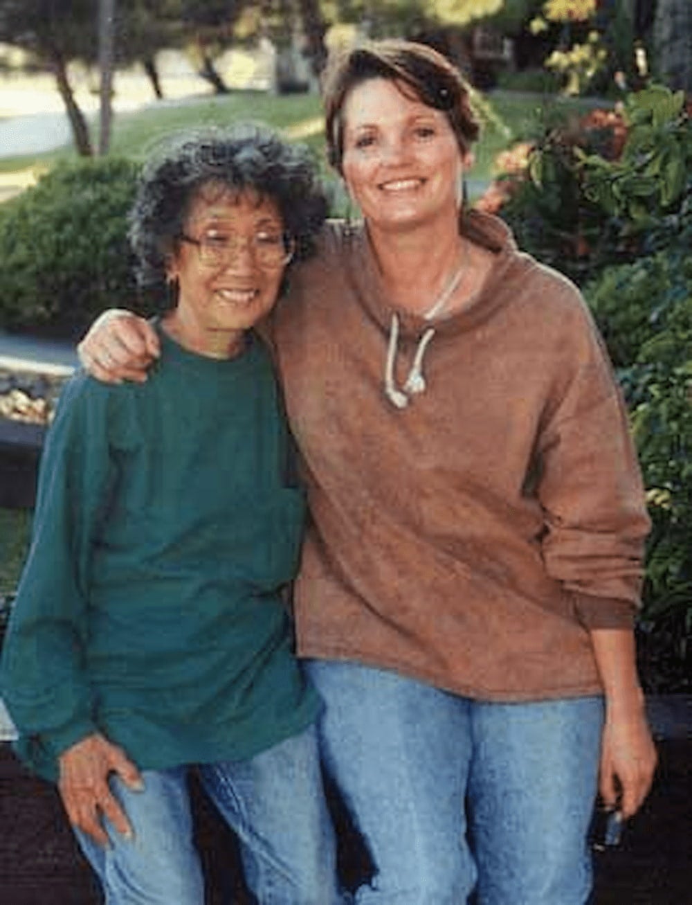 Yuri Kochiyama, wearing jeans and sweater, poses with her arm around Marilyn Buck, also in jeans and sweater. They sit on a wall backed by shrubs.