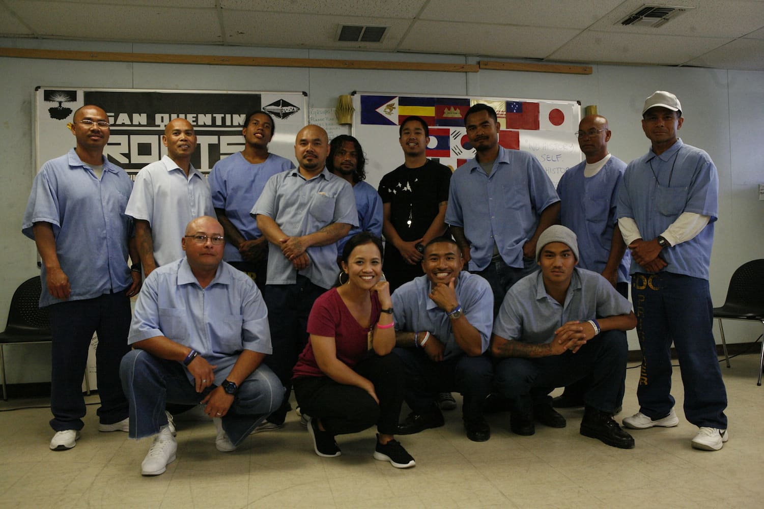 Members of the Asian Prisoner Support Committee pose with eleven inmates in a classroom. One woman and three men crouch in front of a row of nine men.
