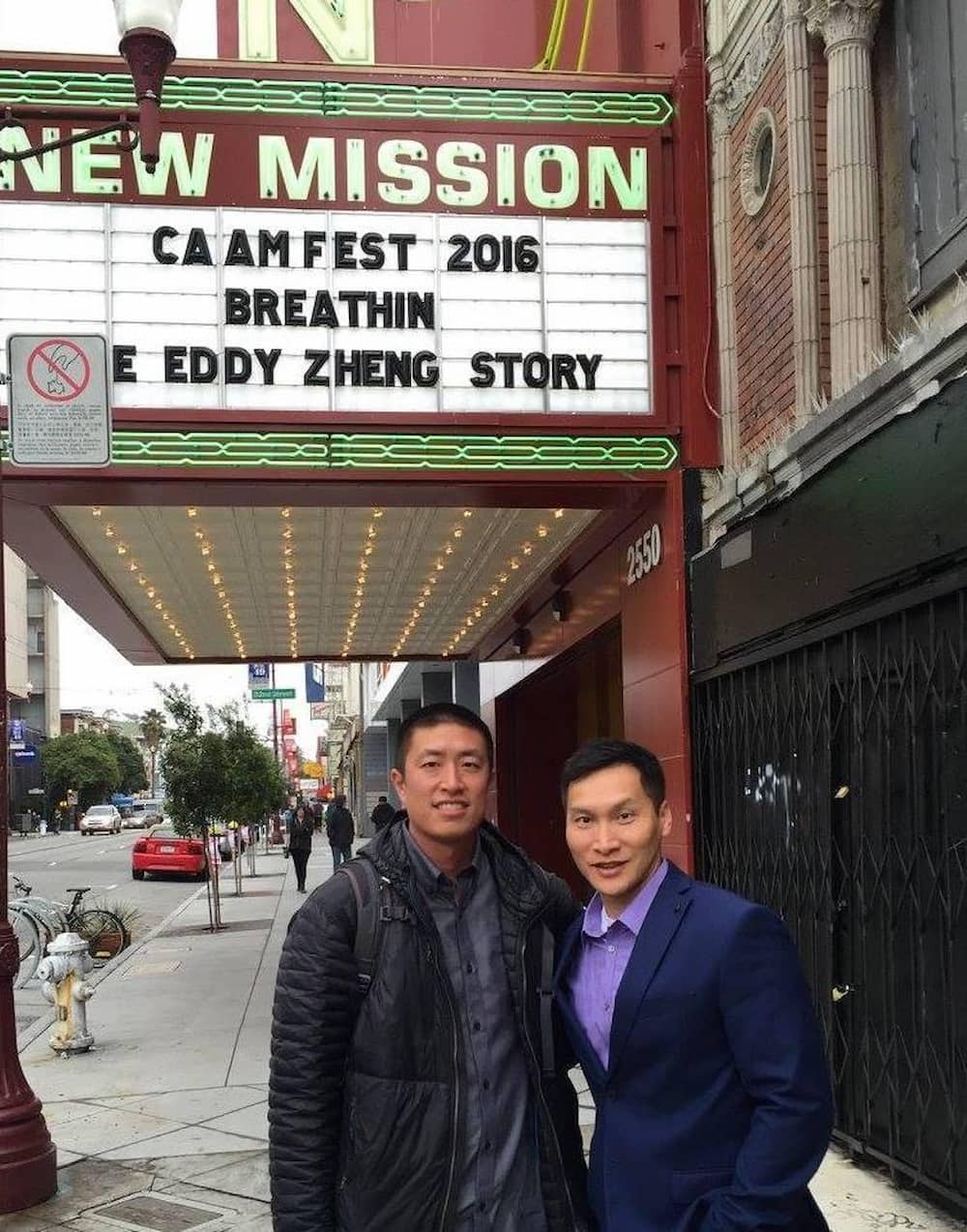 Chinese American Eddy Zheng in a black puffer jacket stands next to director Ben Wang in a suit. They stand in front of marquee promoting their film.