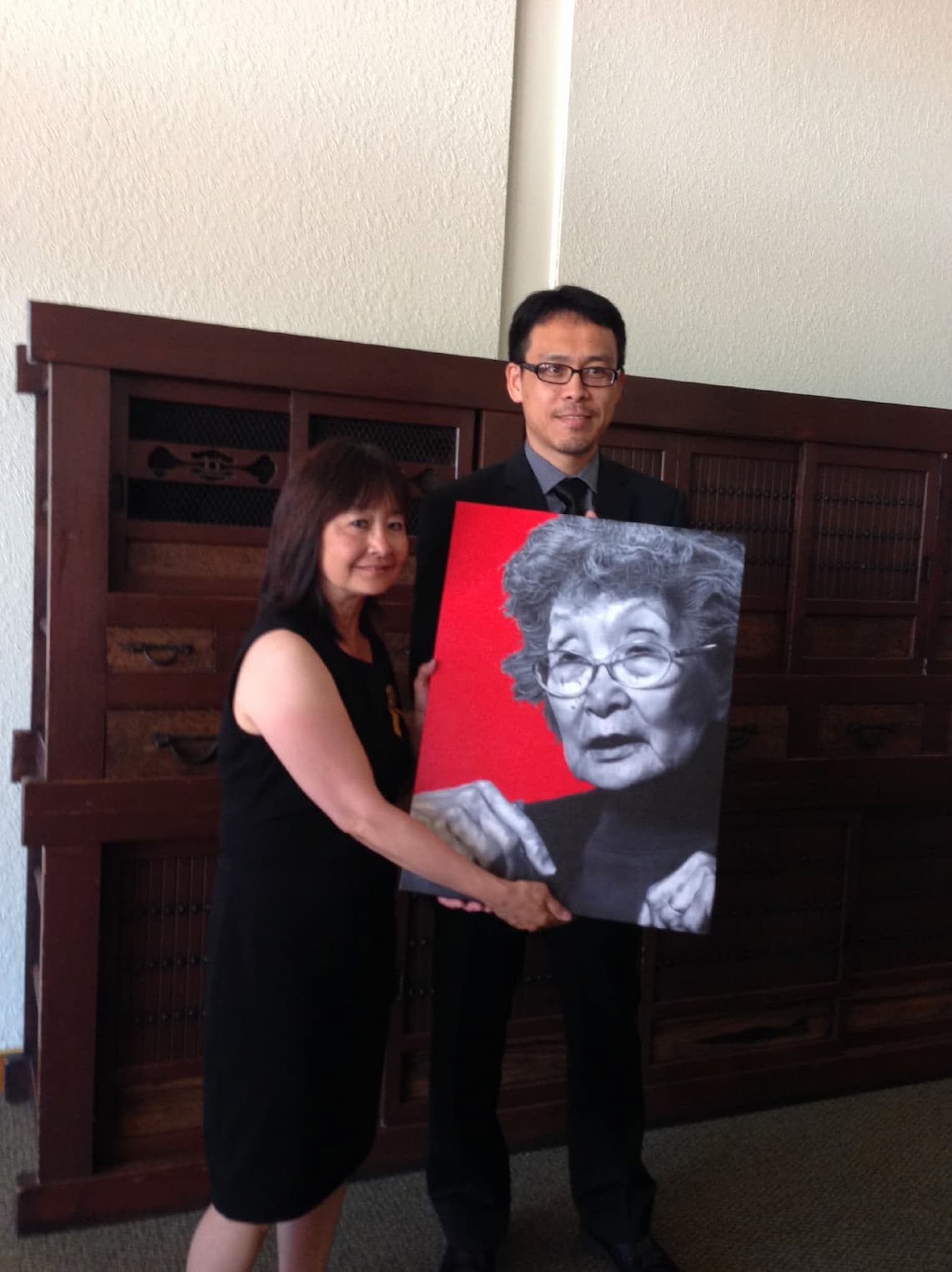 Artist Shuji Nakamura, in a suit, presents his painting of Yuri Kochiyama to Audee Kochiyama-Holman. They pose in front of a wooden cabinet.