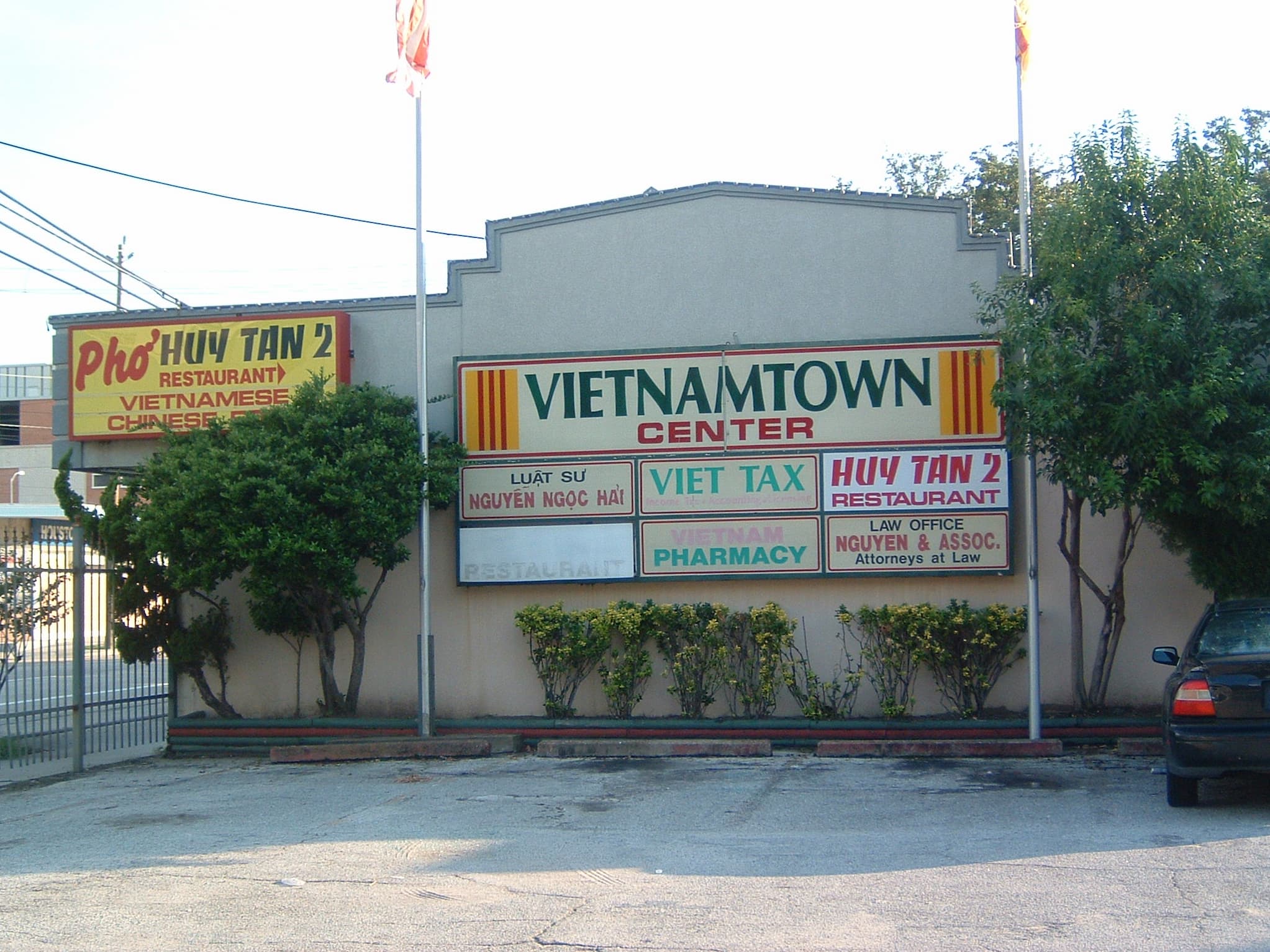 Outdoor strip mall with "Vietnamtown Center" sign on exterior wall and signs for businesses below. Flagpole on either side fly South Vietnamese flags.