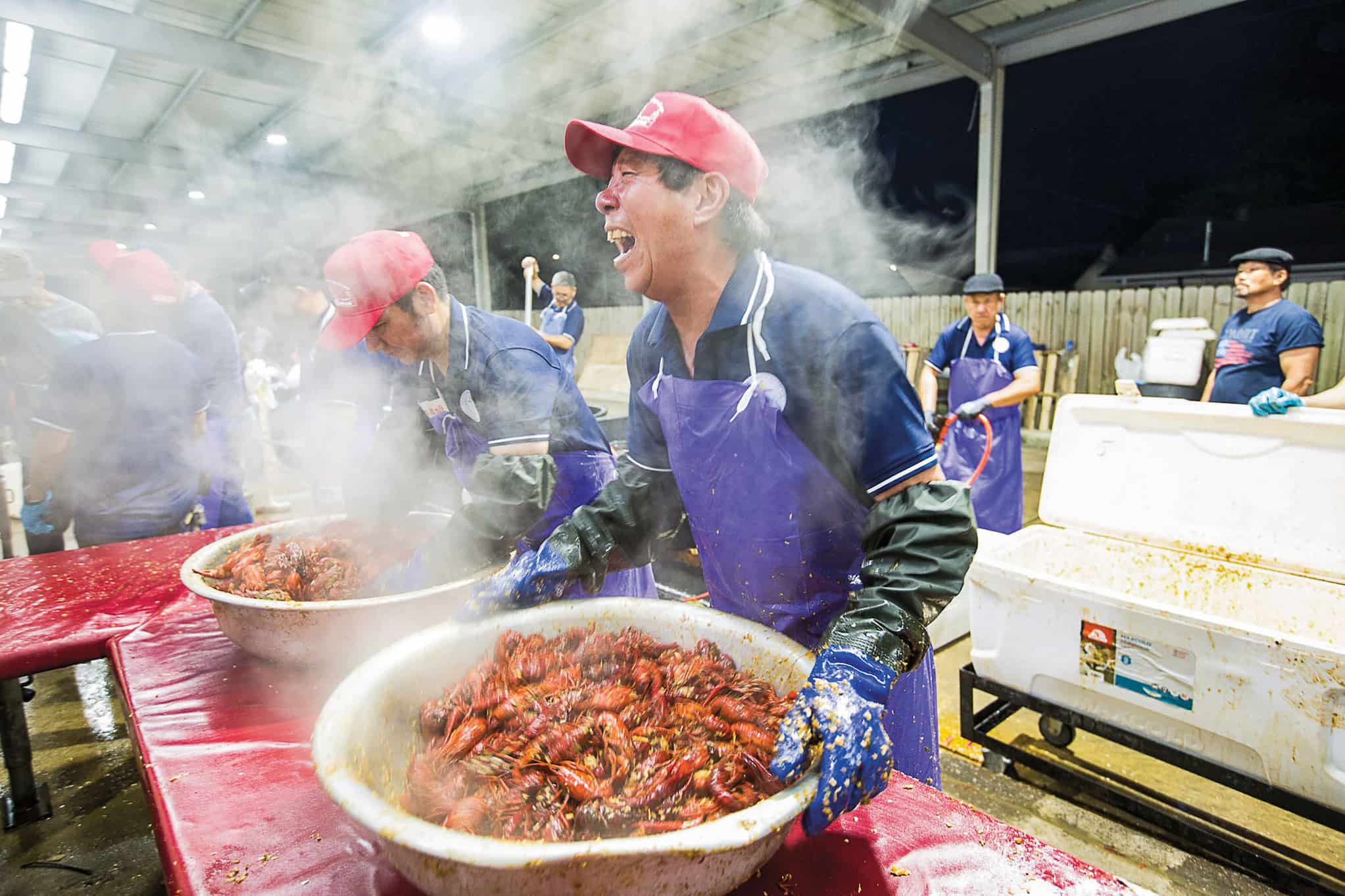 Vietnamese man in an apron, food safety gloves, and a cap laughs as he prepares a large tub of steaming crawfish. Another man works beside him.