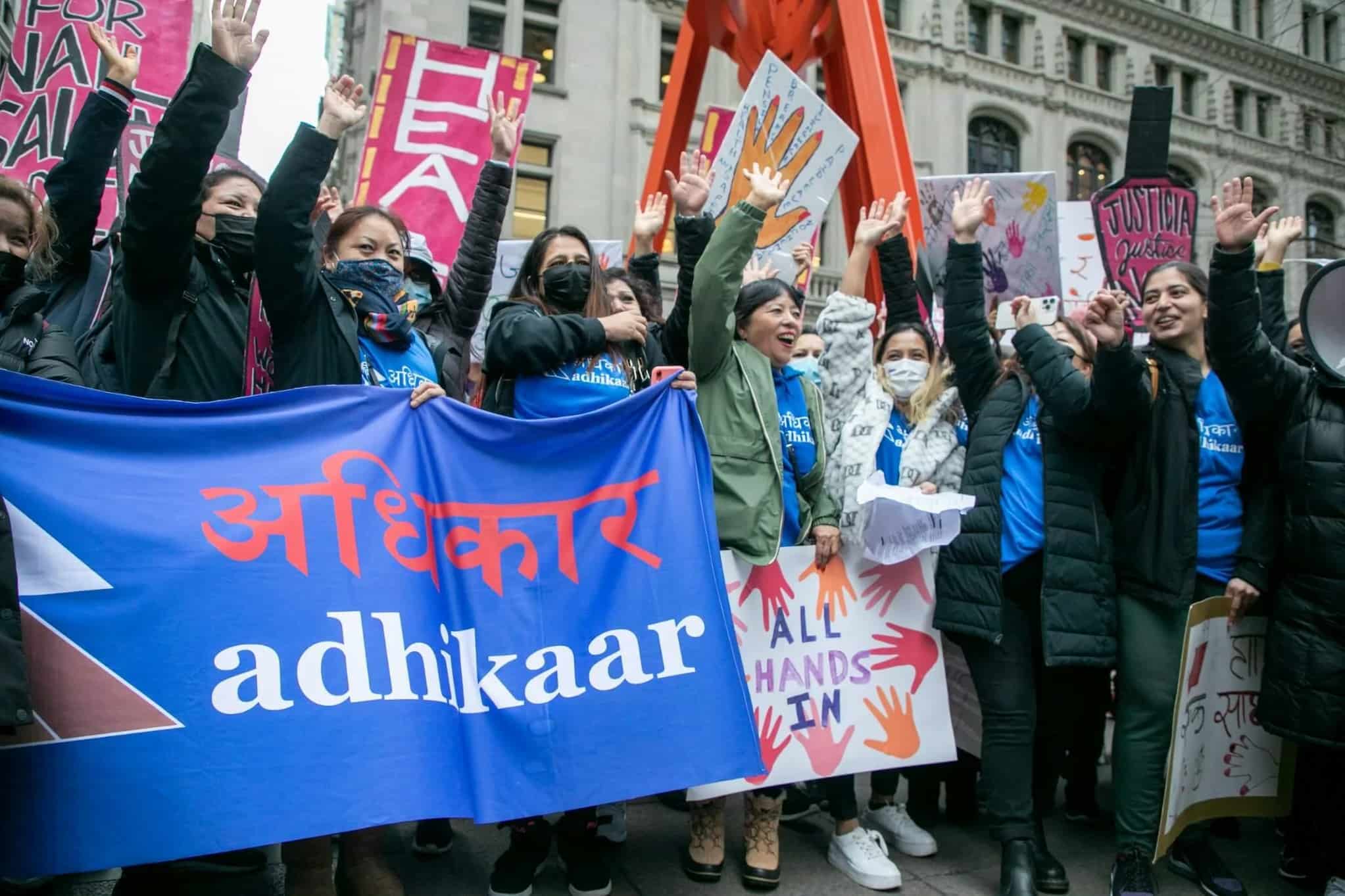 A group of women gather in New York City street with one arm raised above their heads. They stand with signs, including one reading "Adhikaar."