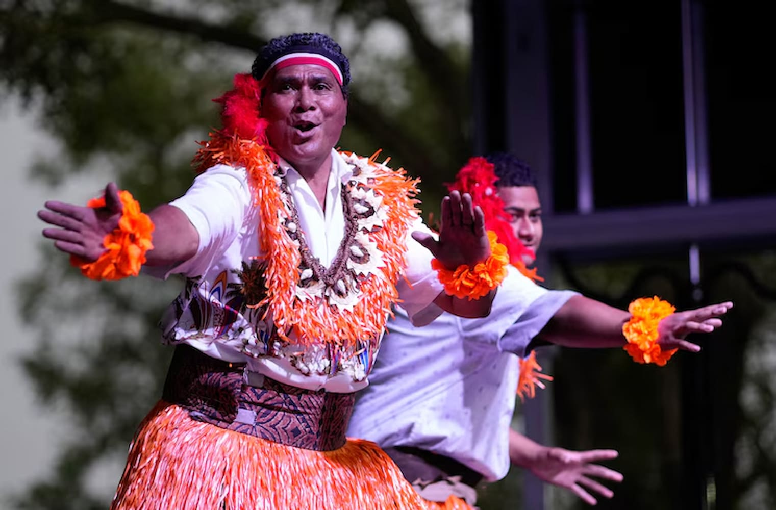 A performer, dressed in a short sleeved shirt, skirt, and floral necklace, performs on stage. Another man, dressed similarly, performs beside him.
