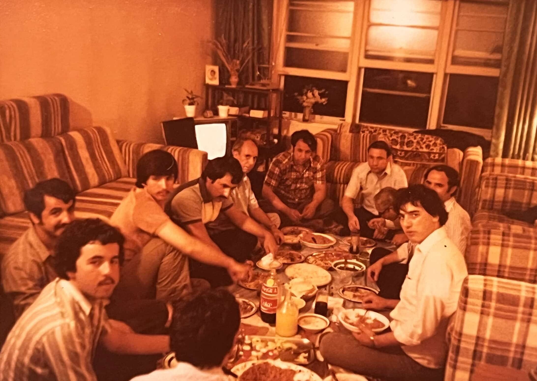 Family and friends of an Afghan migrant, sitting cross-legged on the floor beside sofas and chairs, gather around meal laid out on a tablecloth.