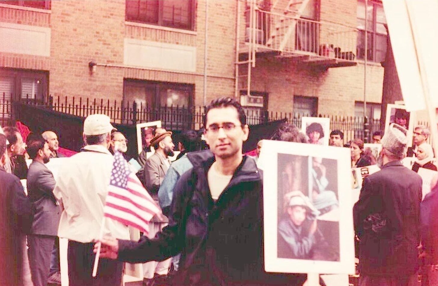 Rameen M. Javid, wearing a jacket and glasses, stands with a U.S. flag in his right hand and a poster held in his left hand during outdoor rally.