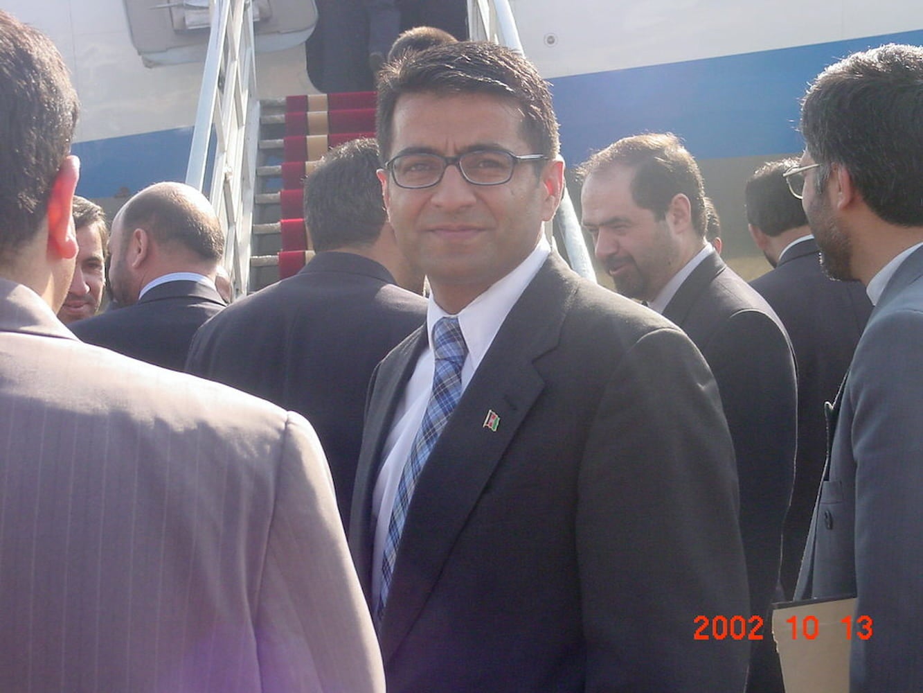 Afghan American Farhad Ahad, with glasses and wearing a suit, stands among similarly dressed men near carpeted boarding stairs of airplane.